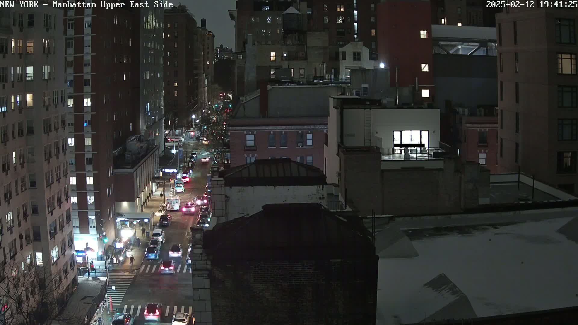A nighttime aerial view of a city street with cars driving on it, surrounded by tall buildings in a snowy urban setting.