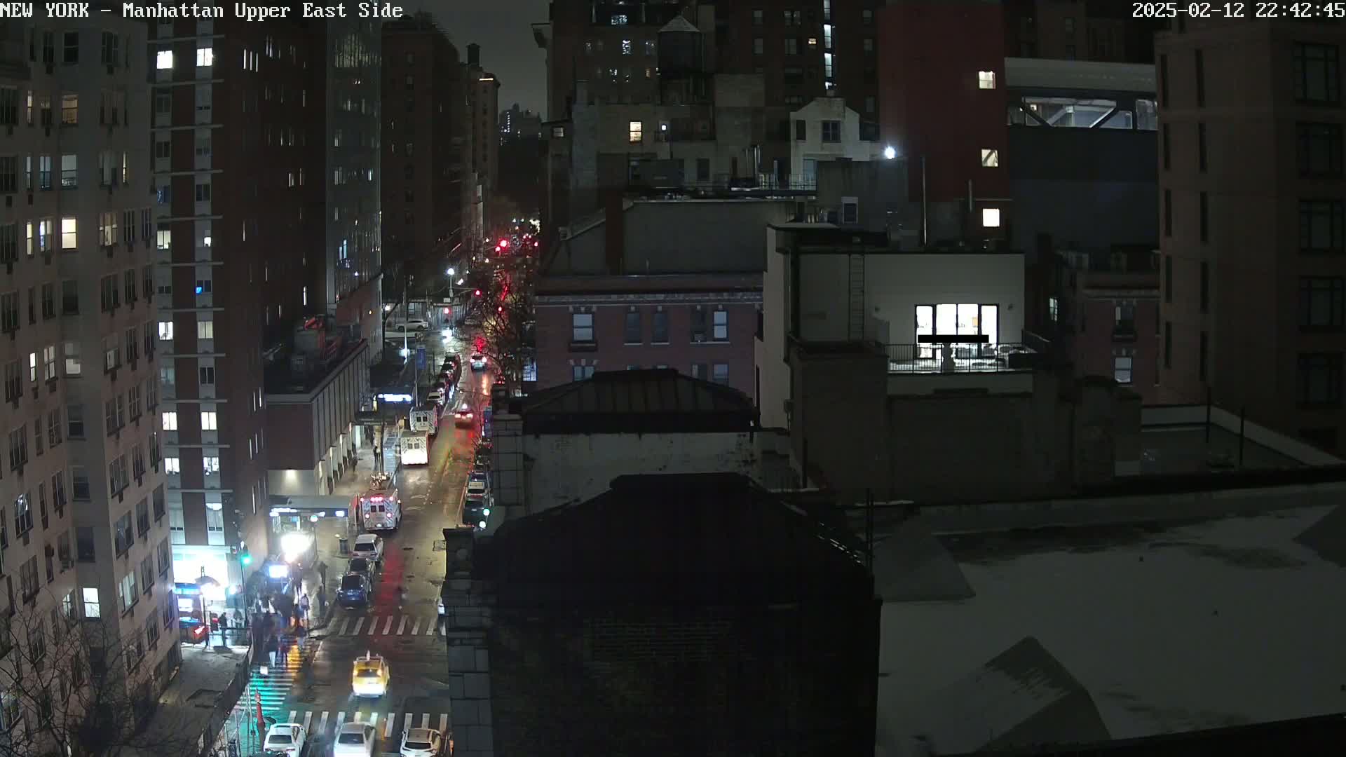 A nighttime aerial view of a city street with traffic, showing several multi-story buildings under a dark, possibly snowy, sky.