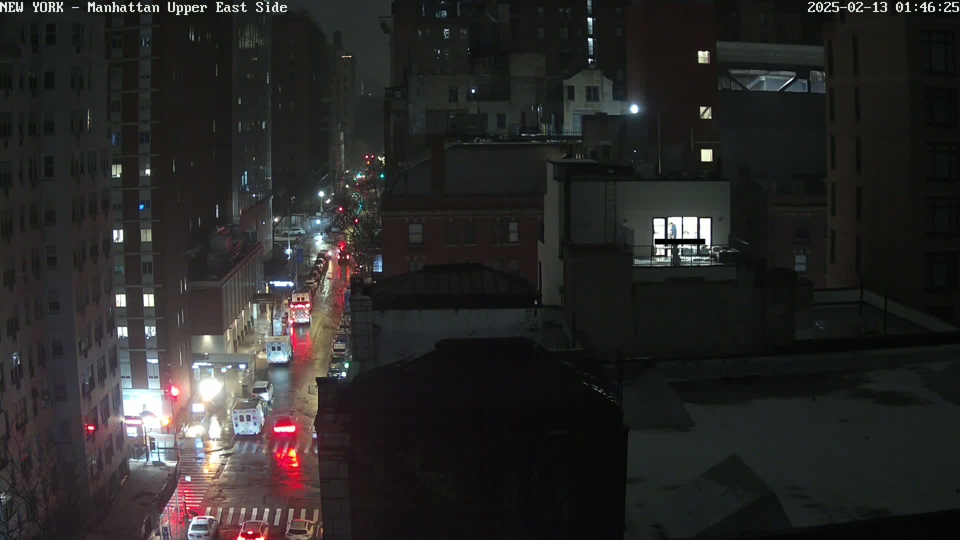 A nighttime, wet city street scene shows several vehicles, including emergency vehicles, moving along a road lined with tall buildings under dim lighting.