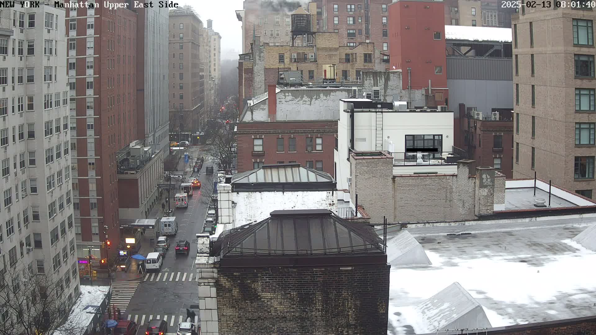 A high-angle, long shot of a city street with light traffic on a cloudy day, showing several multi-story buildings with some snow on their rooftops.