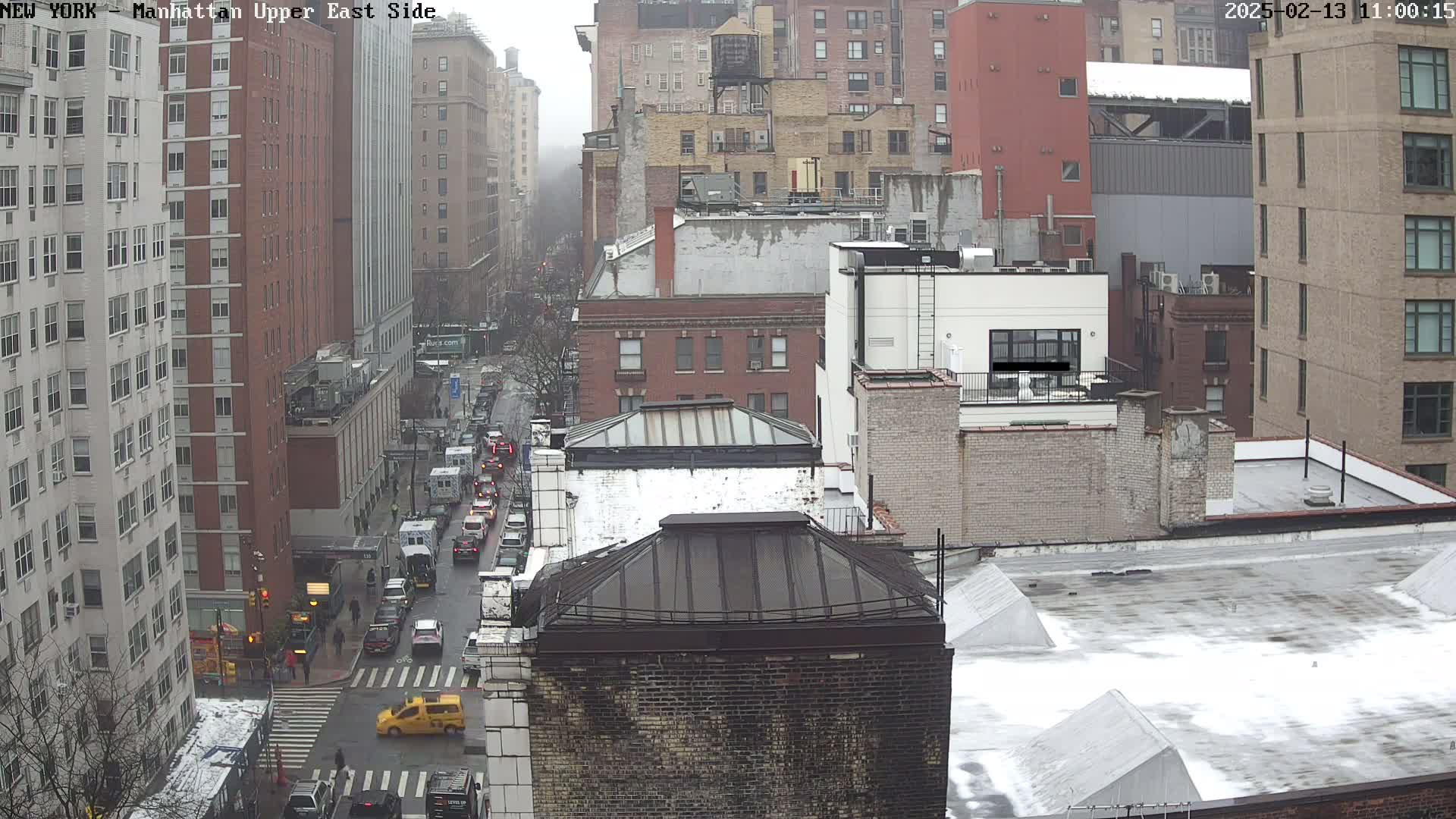 A city street scene on an overcast day shows a view of multiple brick buildings, traffic, and a light dusting of snow on the roofs and ground.