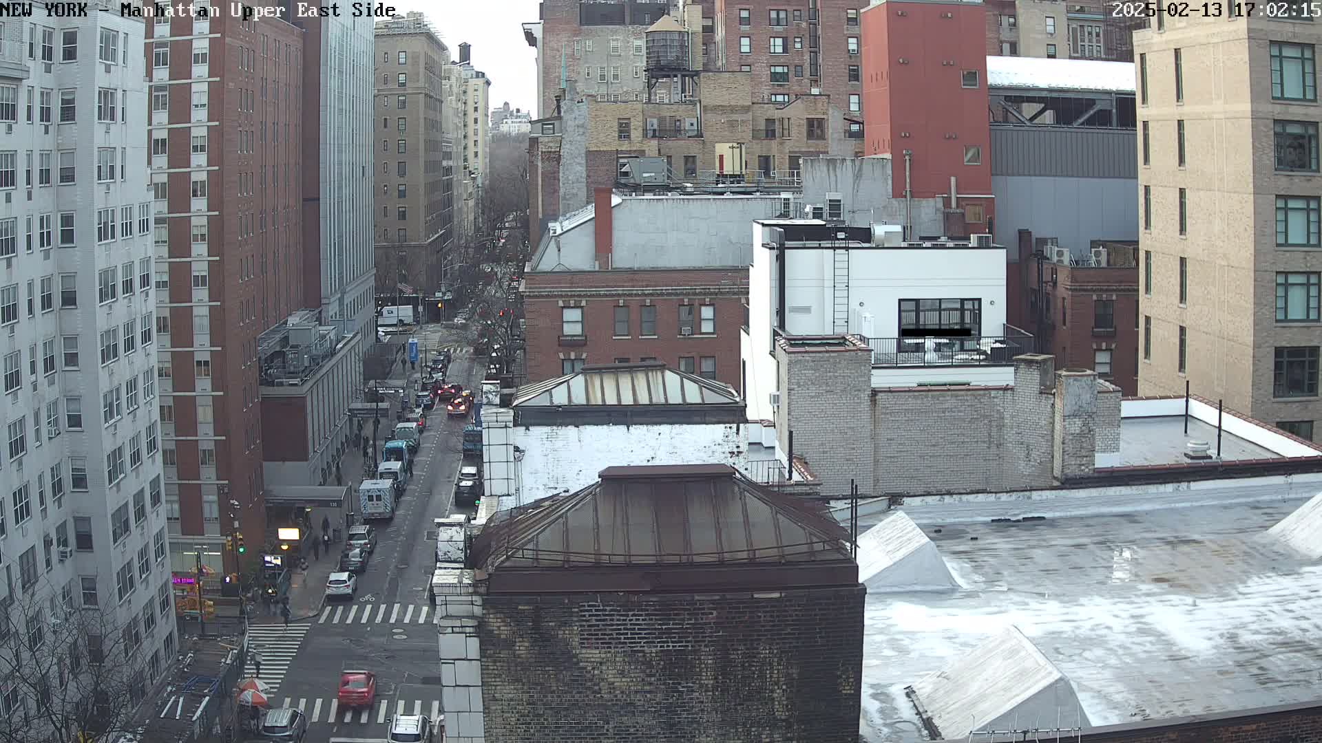 An aerial view of a city street scene with several multi-story buildings, some cars on the street, and a mostly overcast sky.