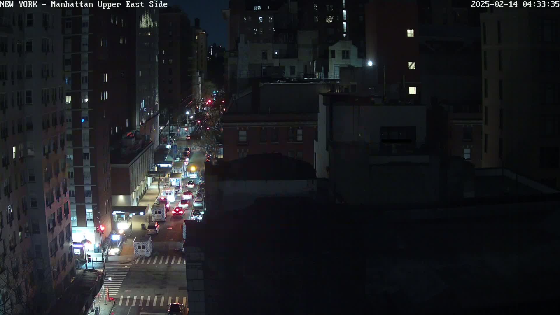 A nighttime, high-angle view of a city street with several cars driving on it, surrounded by tall buildings.