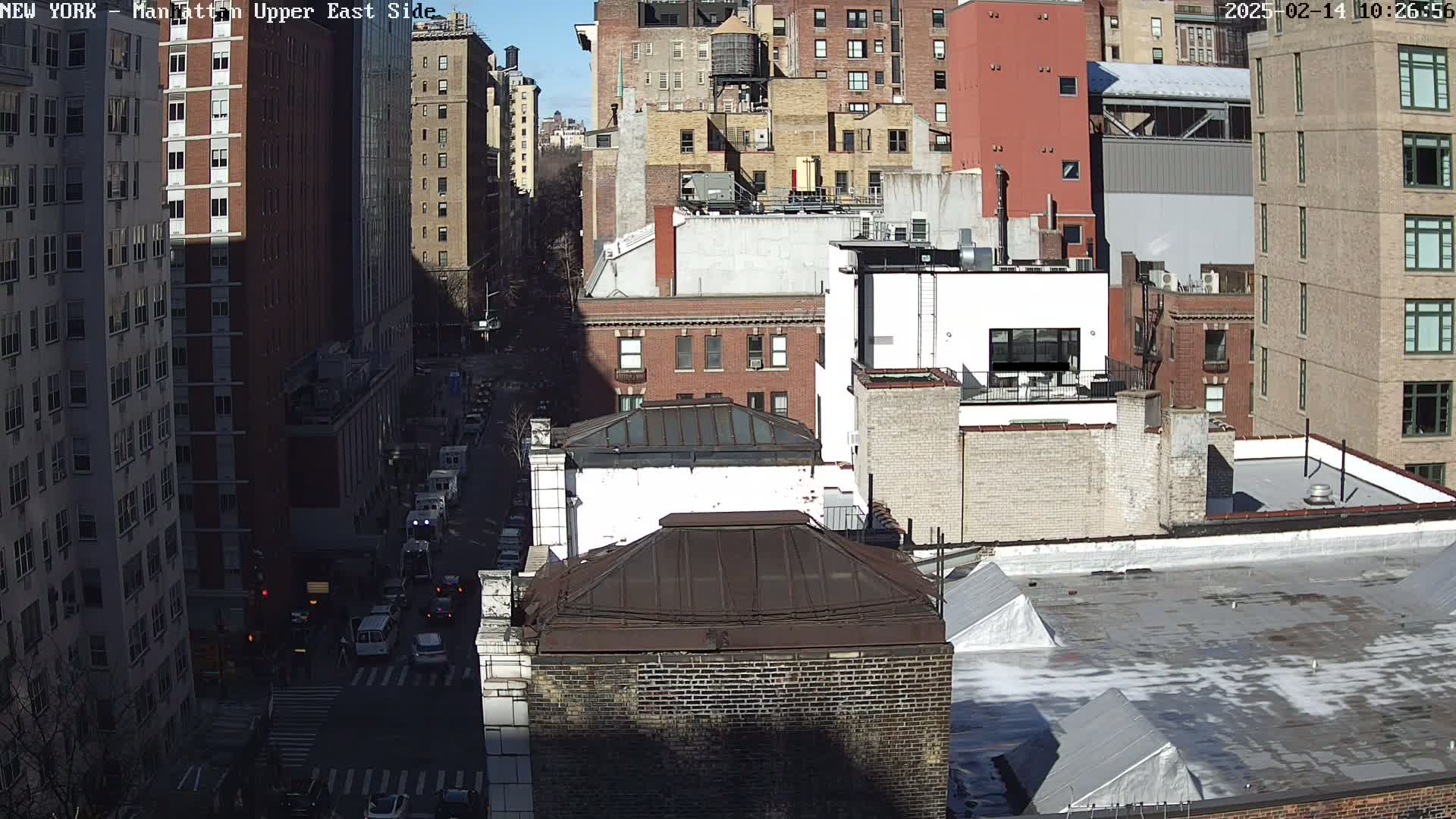 A sunny day view of a city street with several multi-story brick buildings, a few vehicles on the street below, and some light snow or ice on the rooftops.