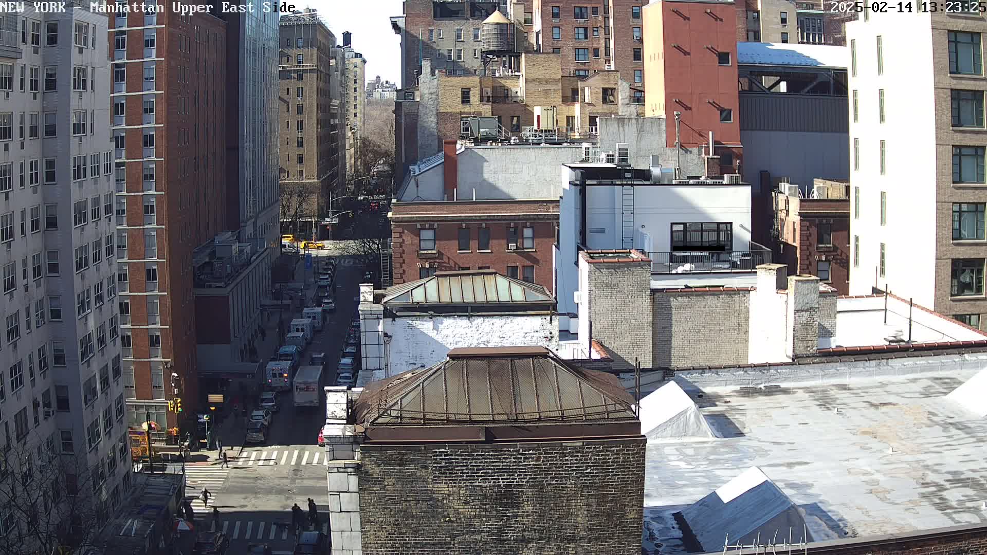 A sunny day view of a city street scene showing a variety of brick buildings, some with rooftop structures, and vehicles on the street below.