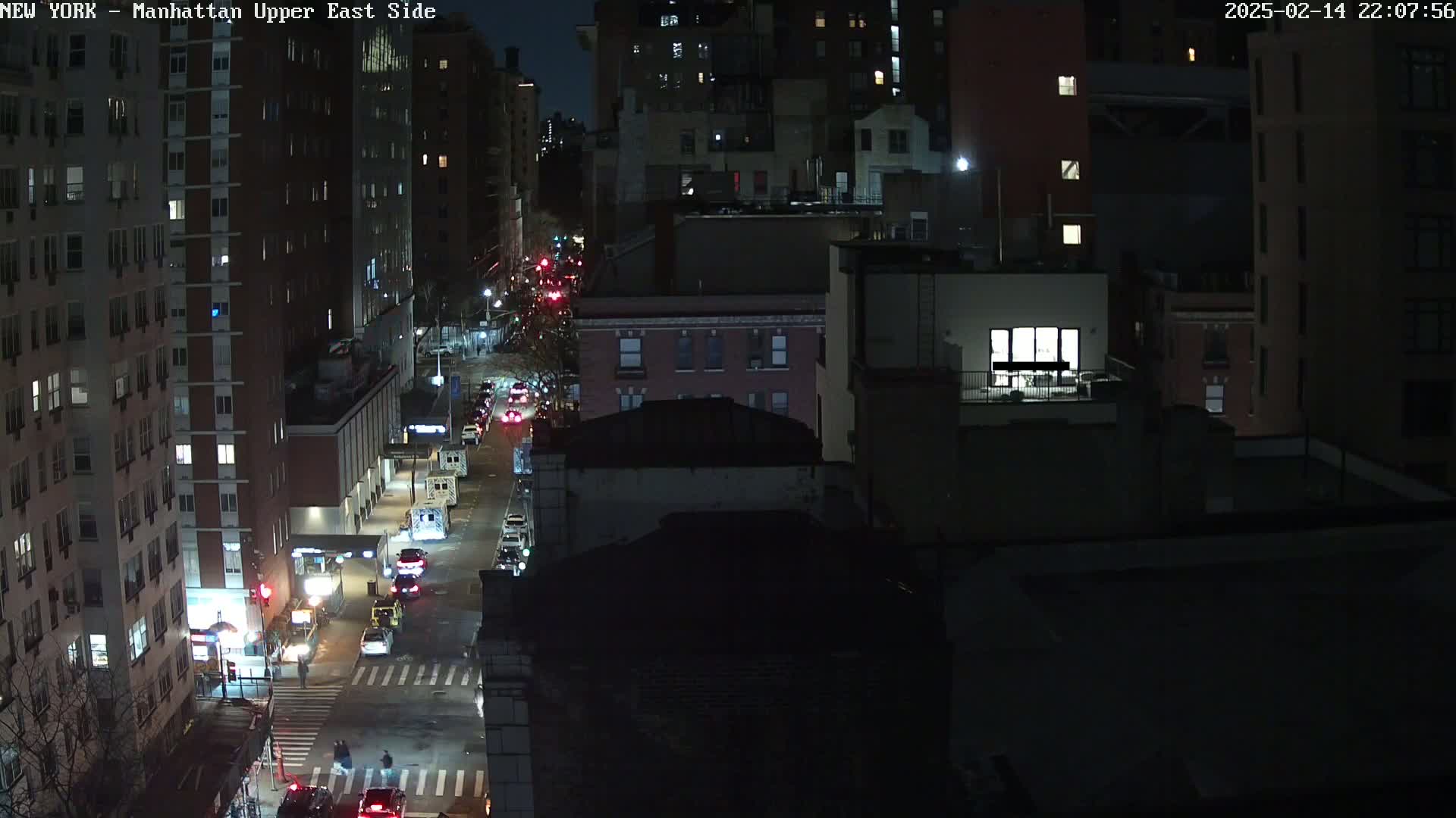 A nighttime, high-angle view of a city street with cars driving on it and buildings lining both sides, under a clear, dark sky.