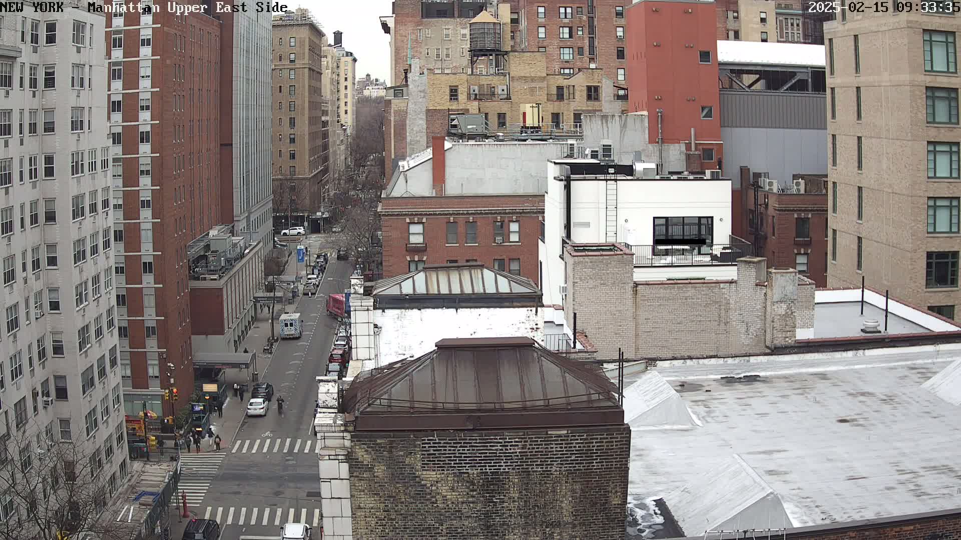 An overcast day reveals a high-angle view of a city street, lined with multi-story brick buildings, and some cars driving on it.