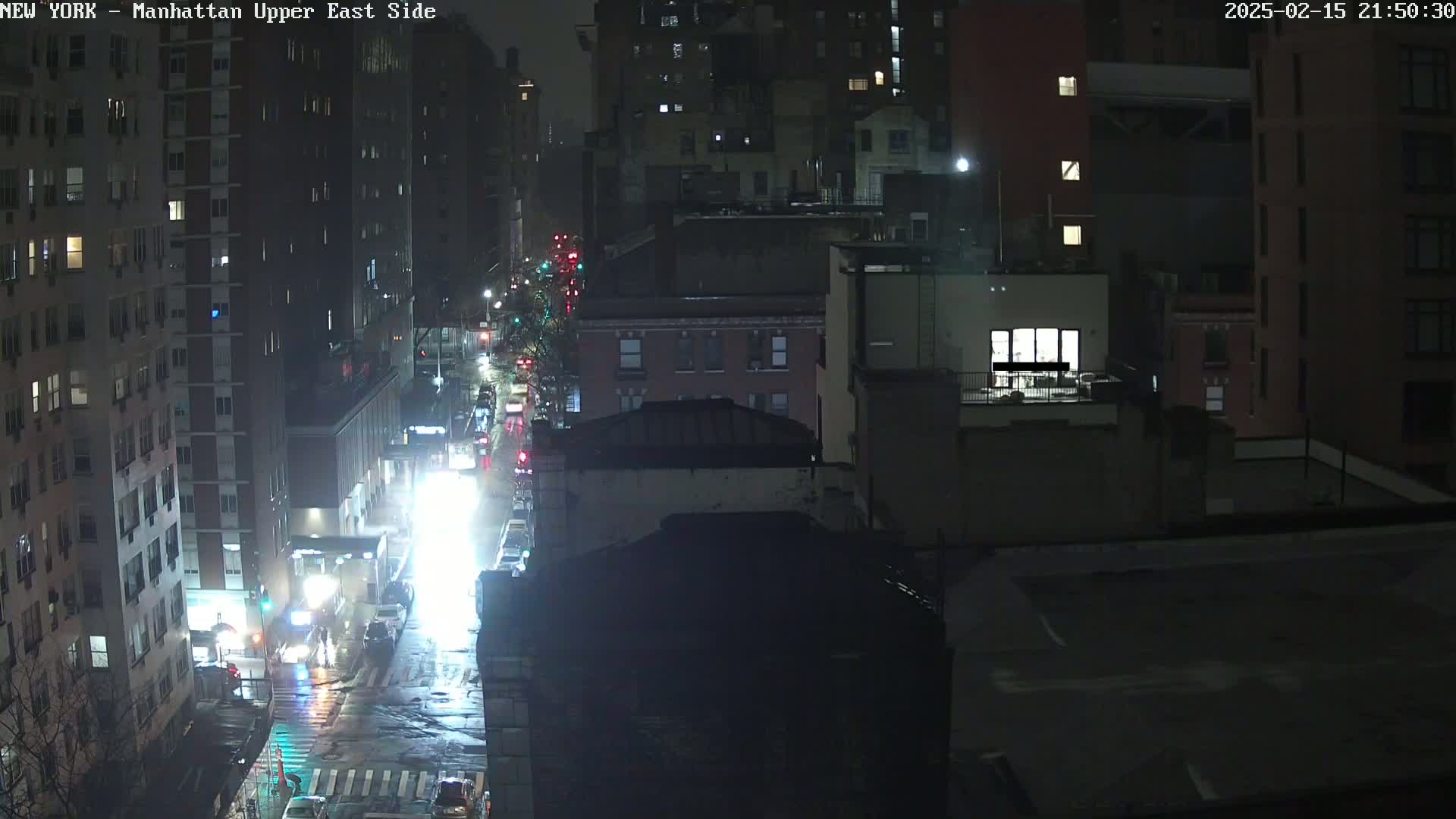 A rain-slicked city street at night, showing traffic moving along a multi-lane road between tall buildings.