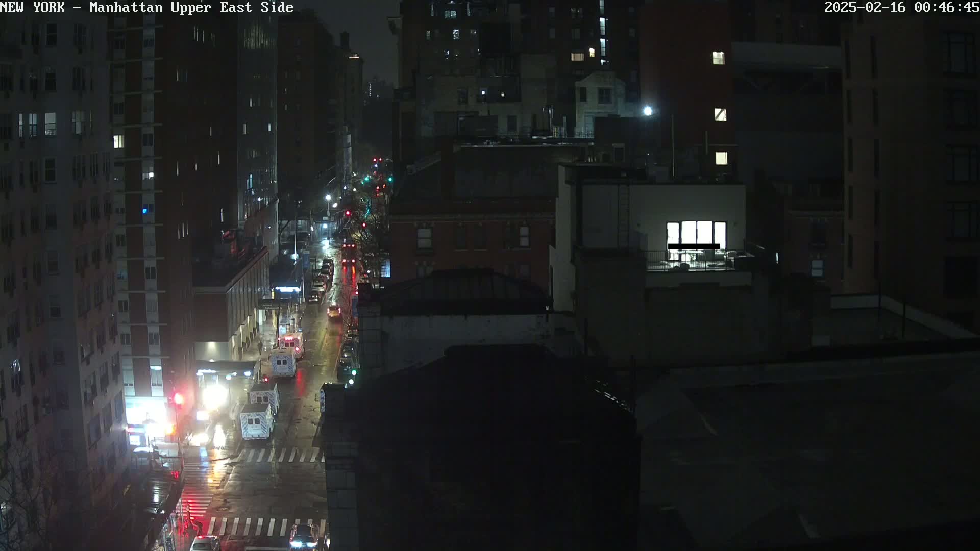 A rain-slicked city street at night, viewed from above, shows several buildings and sparse traffic under dim streetlights.