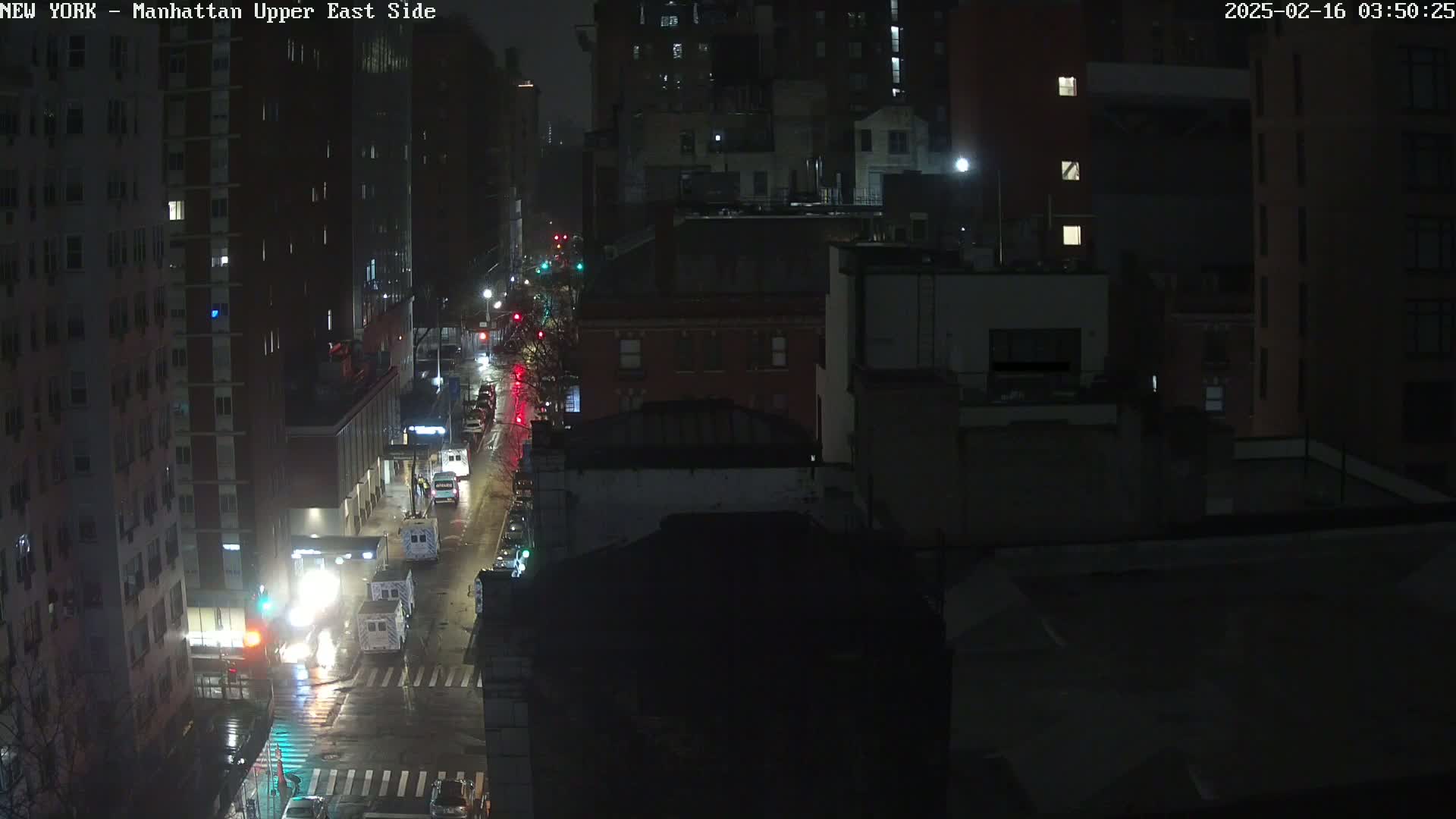 A rain-slicked city street at night shows several vehicles and buildings under dim lighting.