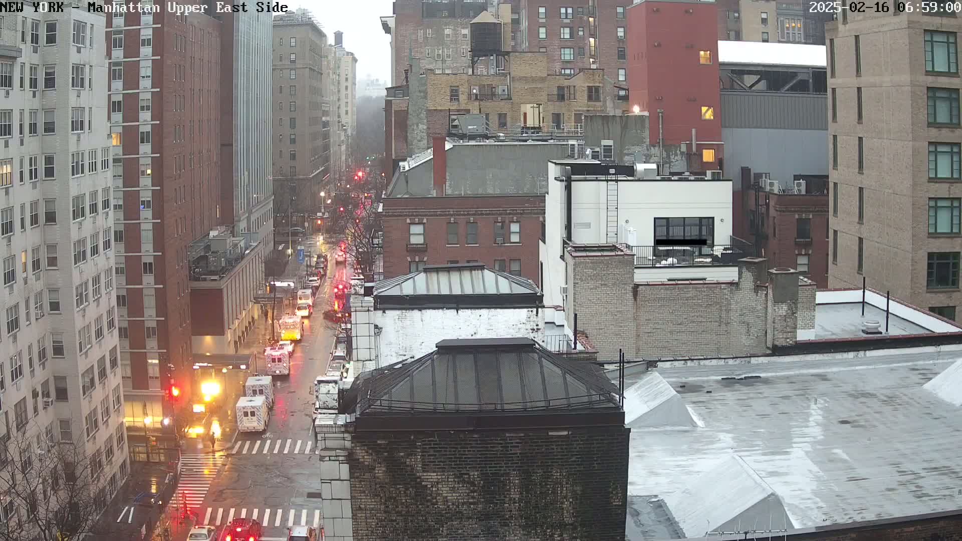 A rainy city street scene shows several buildings, traffic, and emergency vehicles at an intersection.