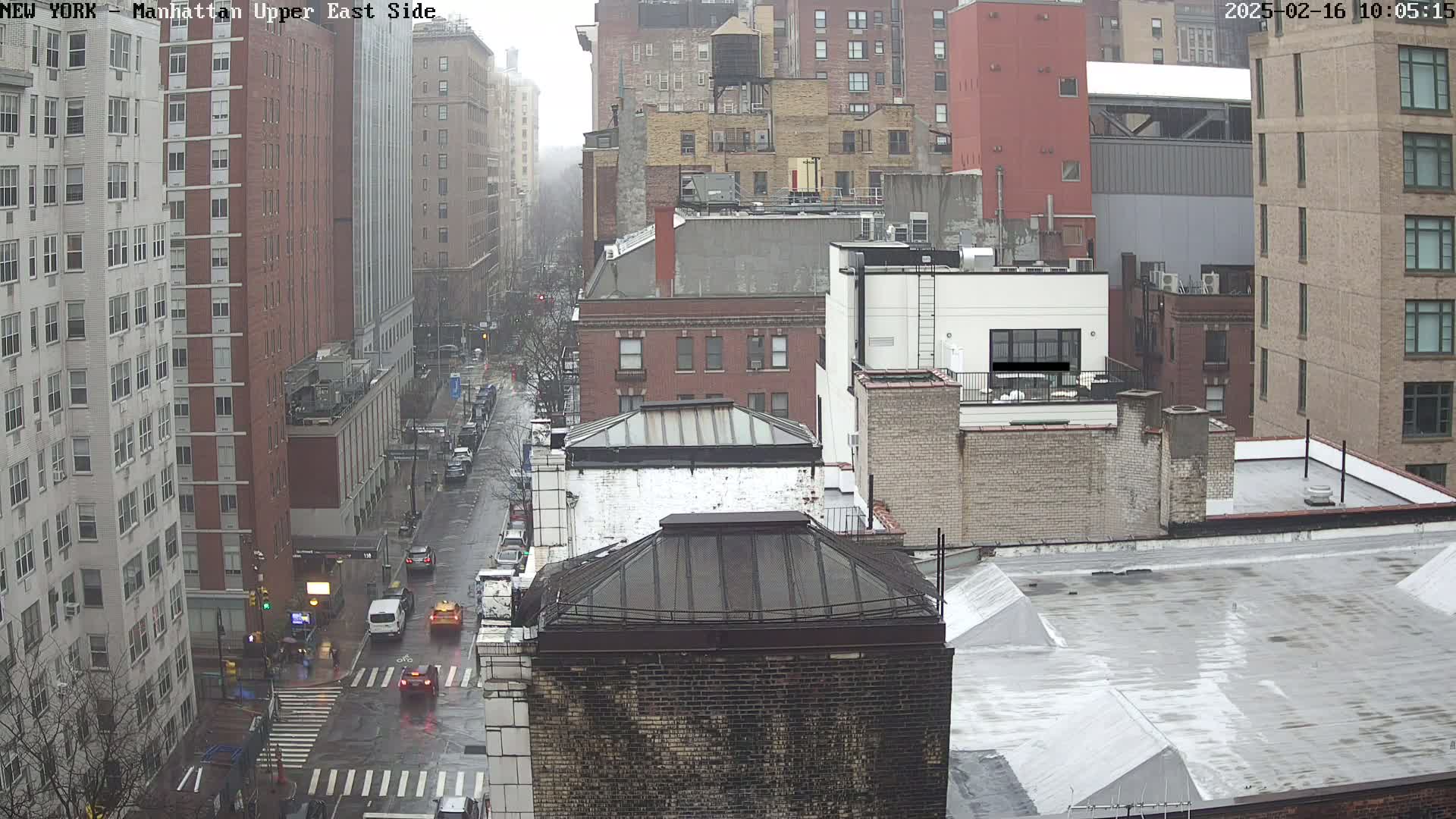 A rainy view of a city street, showing several multi-story buildings and a few cars driving on a wet road.