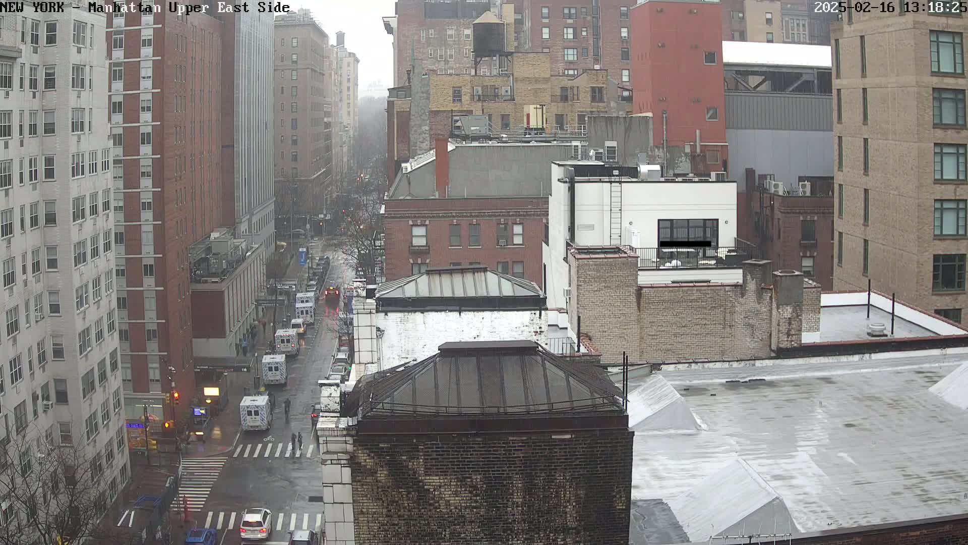A rain-slicked city street scene viewed from above shows several multi-story buildings, with several emergency vehicles parked along the street.