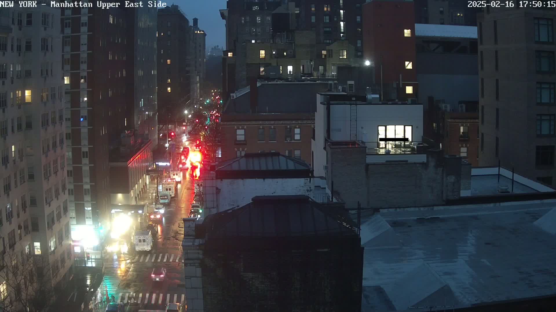 A rain-slicked city street at night is filled with traffic, including emergency vehicles, as seen from an elevated vantage point overlooking several multi-story buildings.