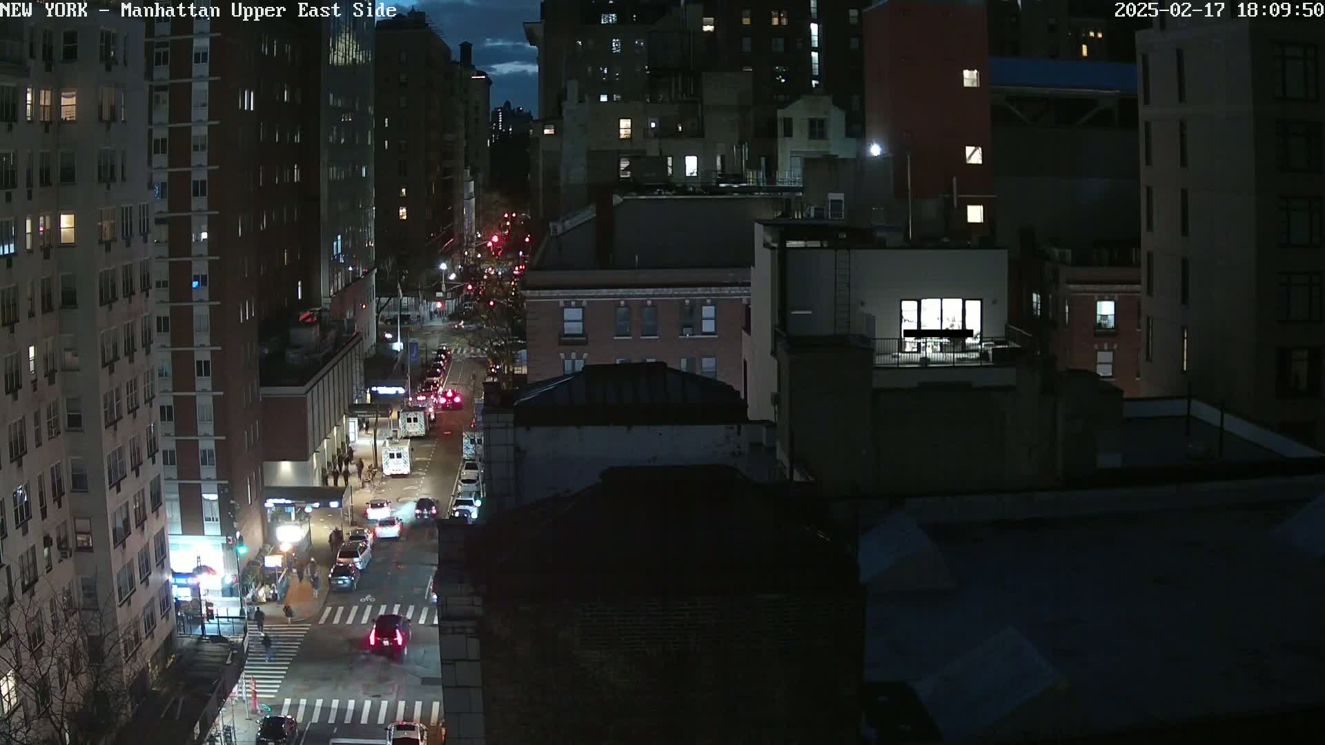 A nighttime aerial view of a city street with cars driving and pedestrians walking, under a partly cloudy sky.