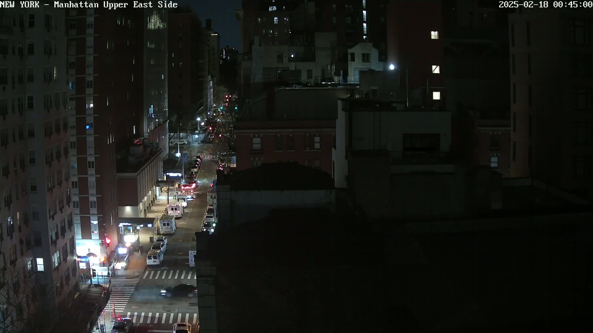 A nighttime aerial view of a city street shows several vehicles, including emergency vehicles, parked and driving along a street lined with dark buildings.