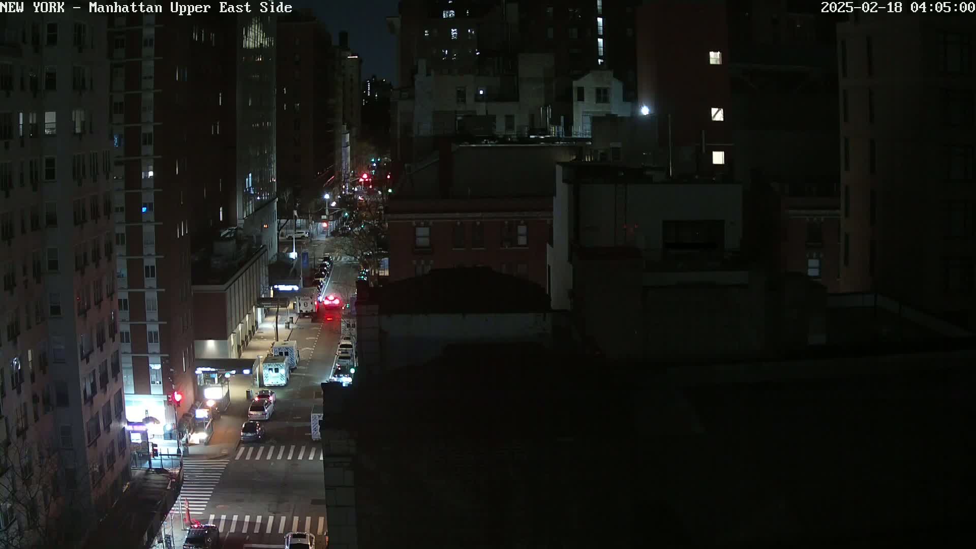 A nighttime, high-angle view of city streets with several vehicles, including emergency vehicles, driving along a roadway between tall buildings.