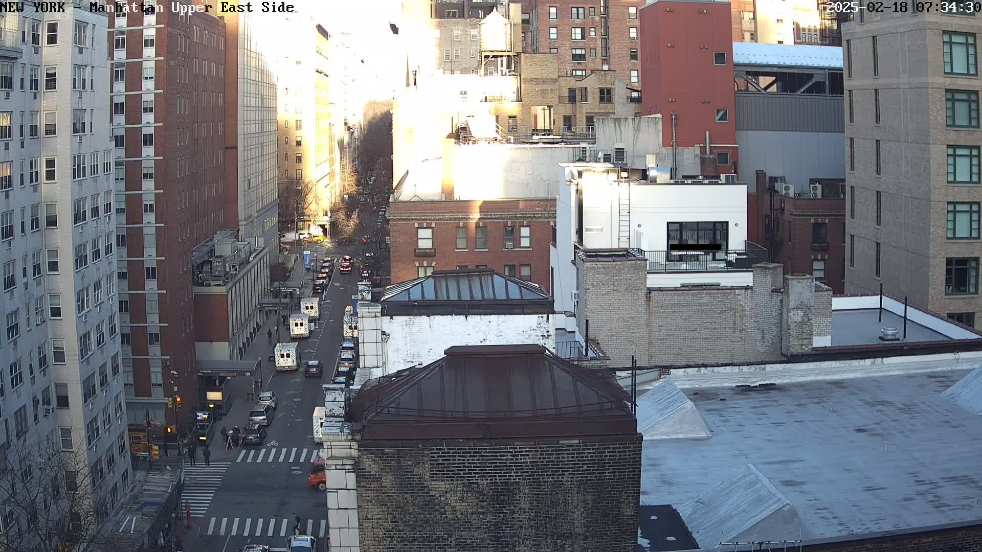 A sunny, city street scene viewed from above shows several multi-story buildings and vehicles on a street below.