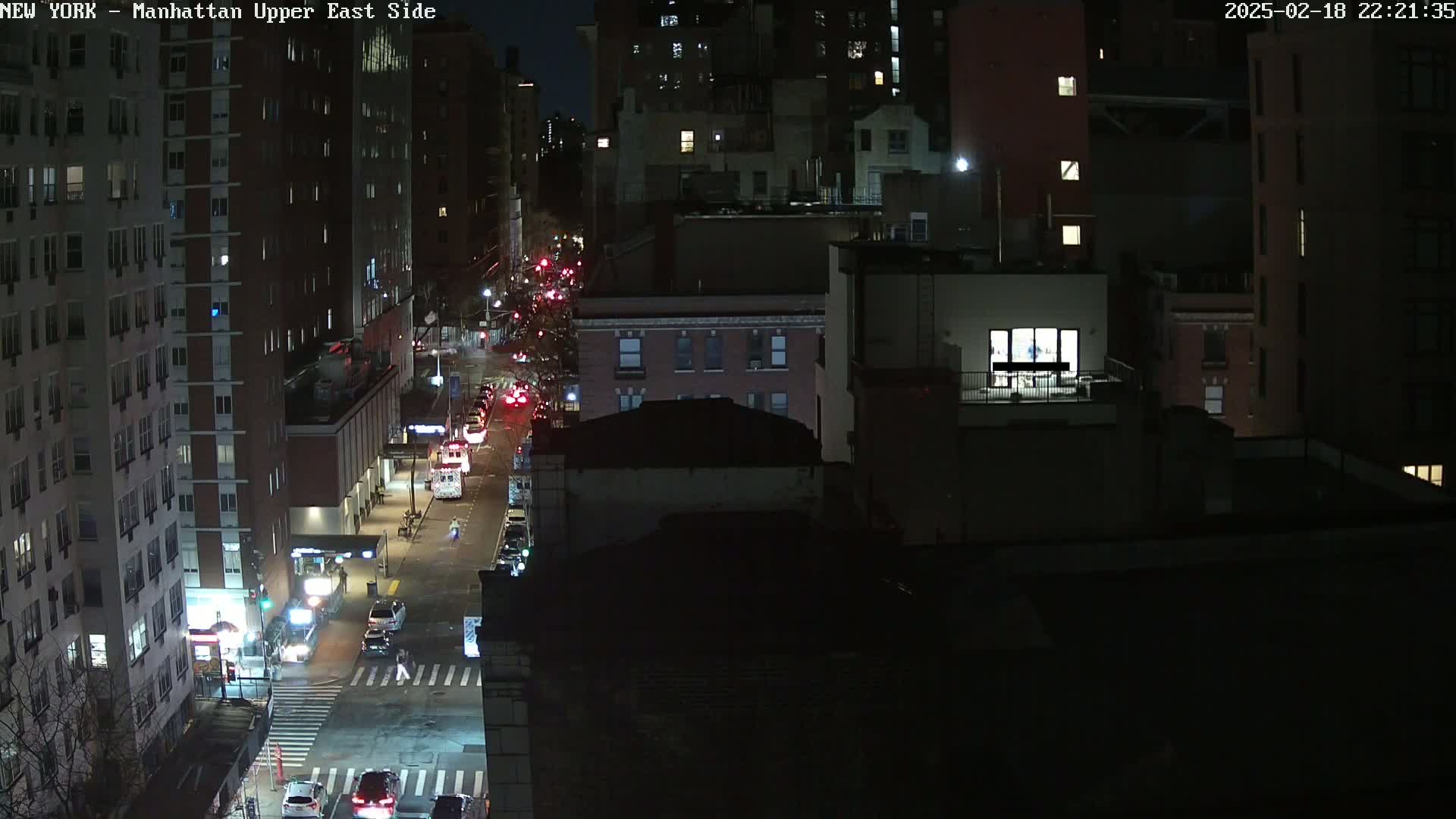 A nighttime aerial view of a city street shows several cars driving on a road between tall buildings under dark conditions.