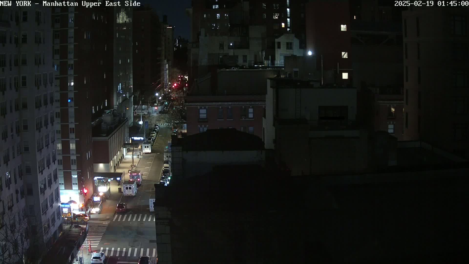 A nighttime aerial view of a city street with several buildings and a few vehicles, including emergency vehicles, under dark conditions.