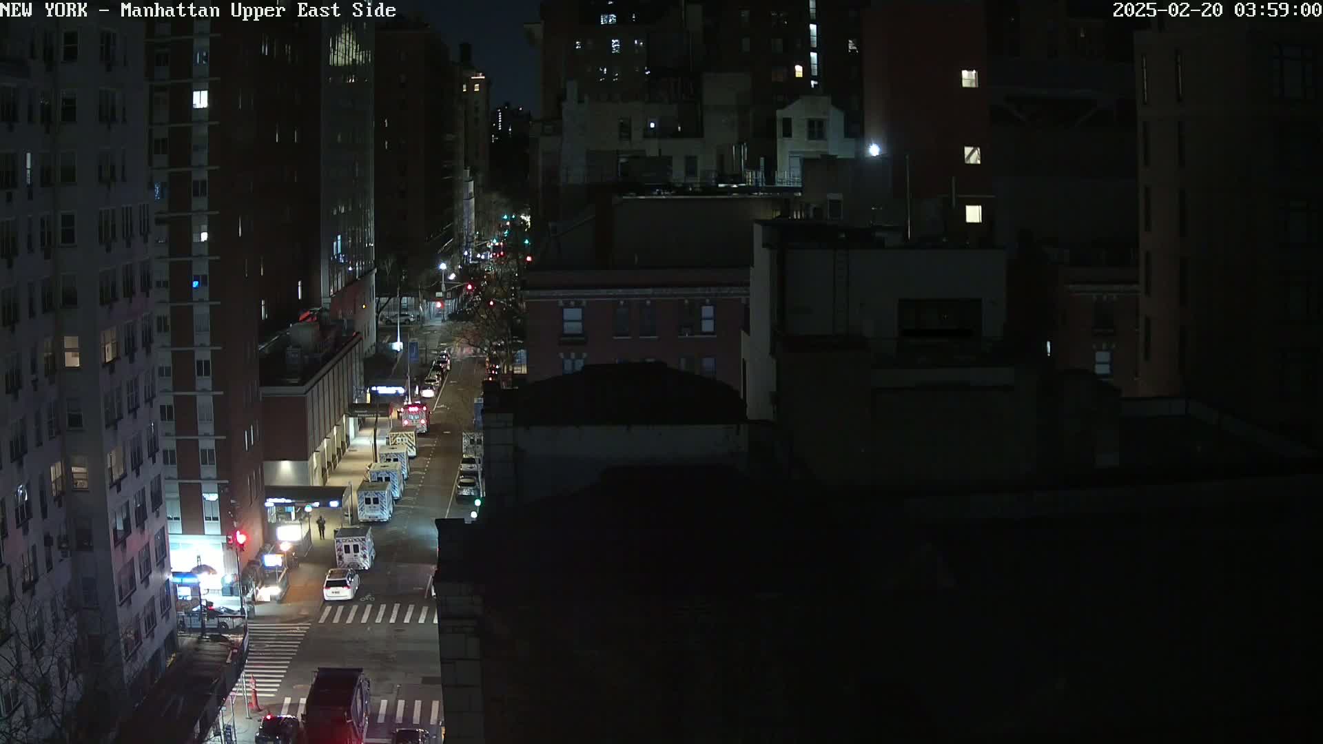 A nighttime, high-angle view of a city street with several vehicles, including what appear to be emergency vehicles, parked along the side of the road, surrounded by dark multi-story buildings.