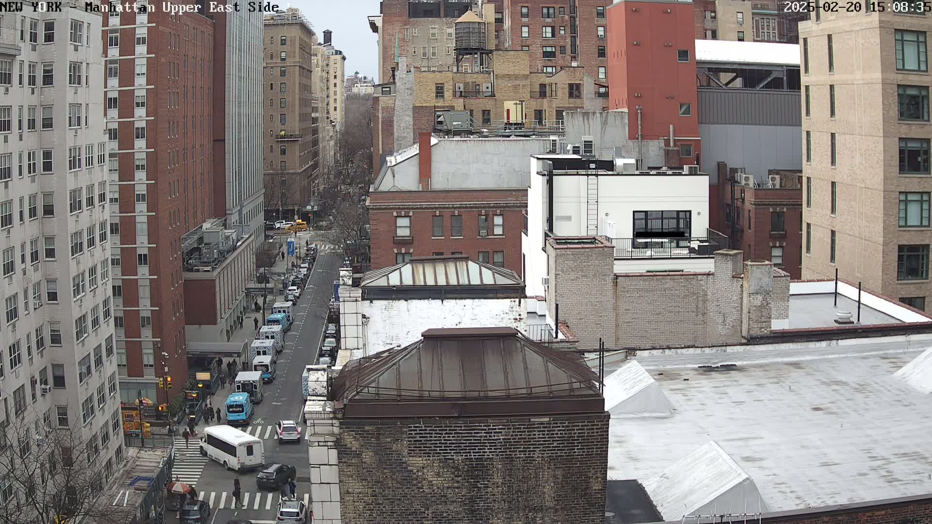 A high-angle view of a city street, on an overcast day, shows several city blocks with multi-story buildings and several vehicles parked along the street and crossing the intersection.