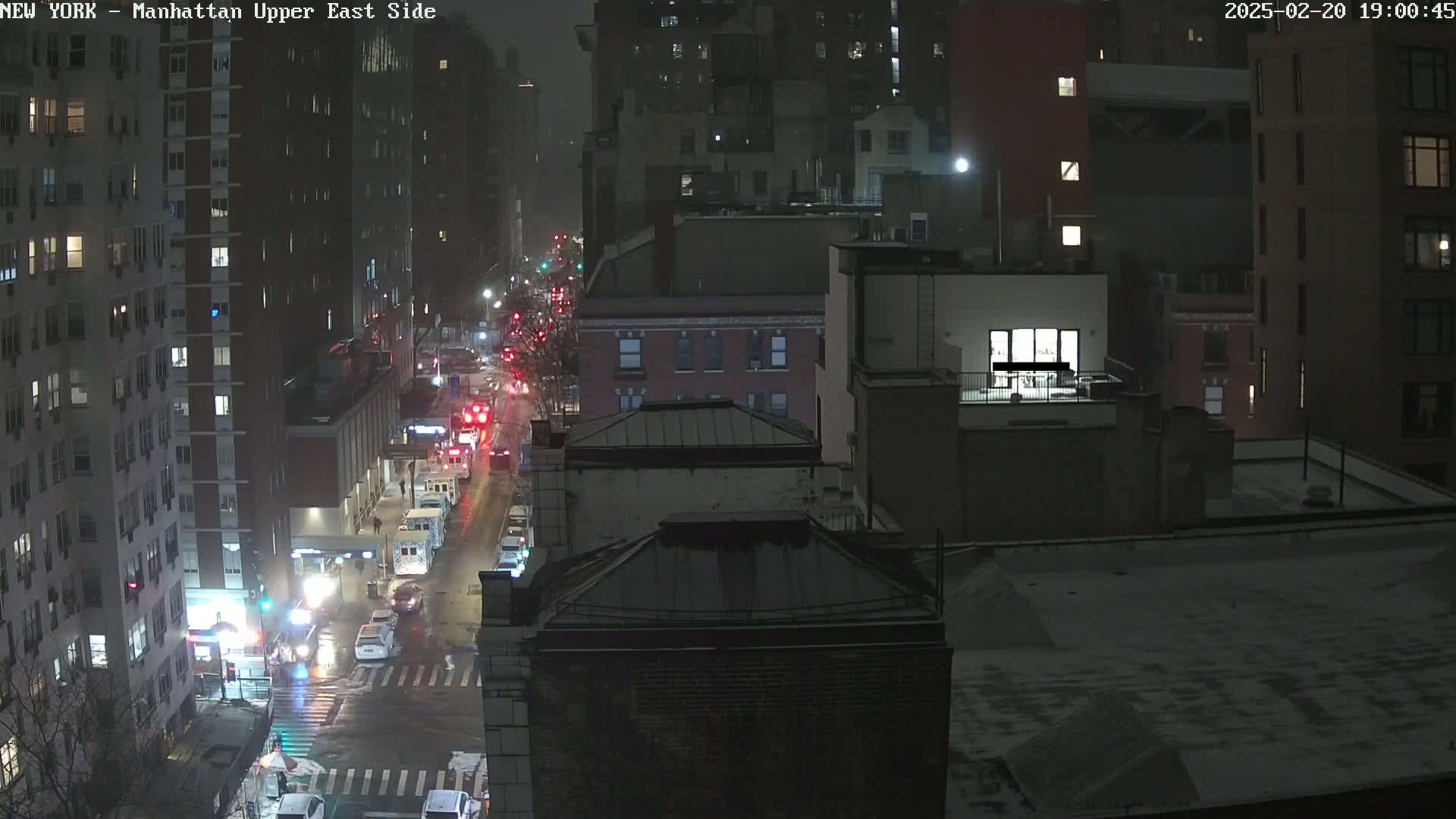 A nighttime, snowy view of a city street with traffic and buildings.