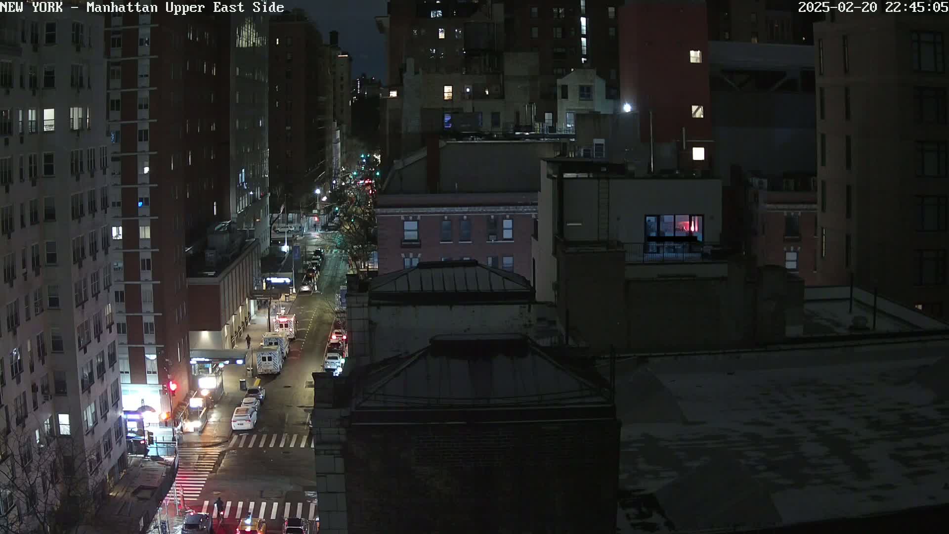 A nighttime, snowy view of a city street with several multi-story buildings and sparse traffic.