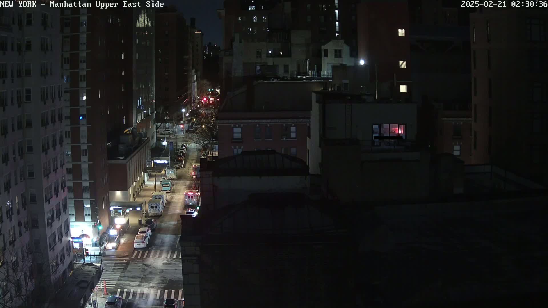 A nighttime, snowy street scene shows several city blocks with tall buildings, some vehicles including emergency vehicles, and lightly trafficked roads.