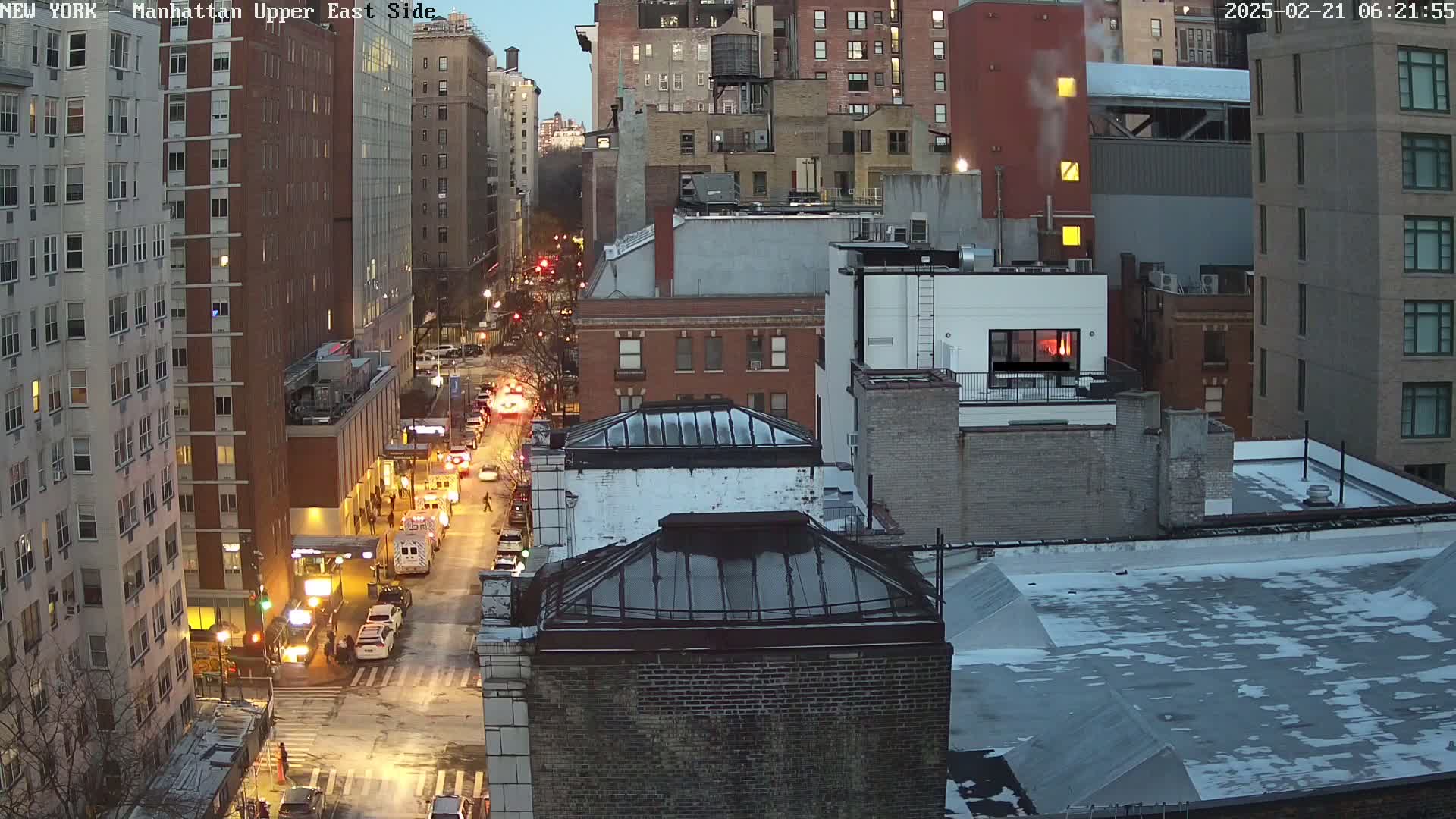 A twilight view of a city street at night, with some snow on the rooftops and cars driving down the road.