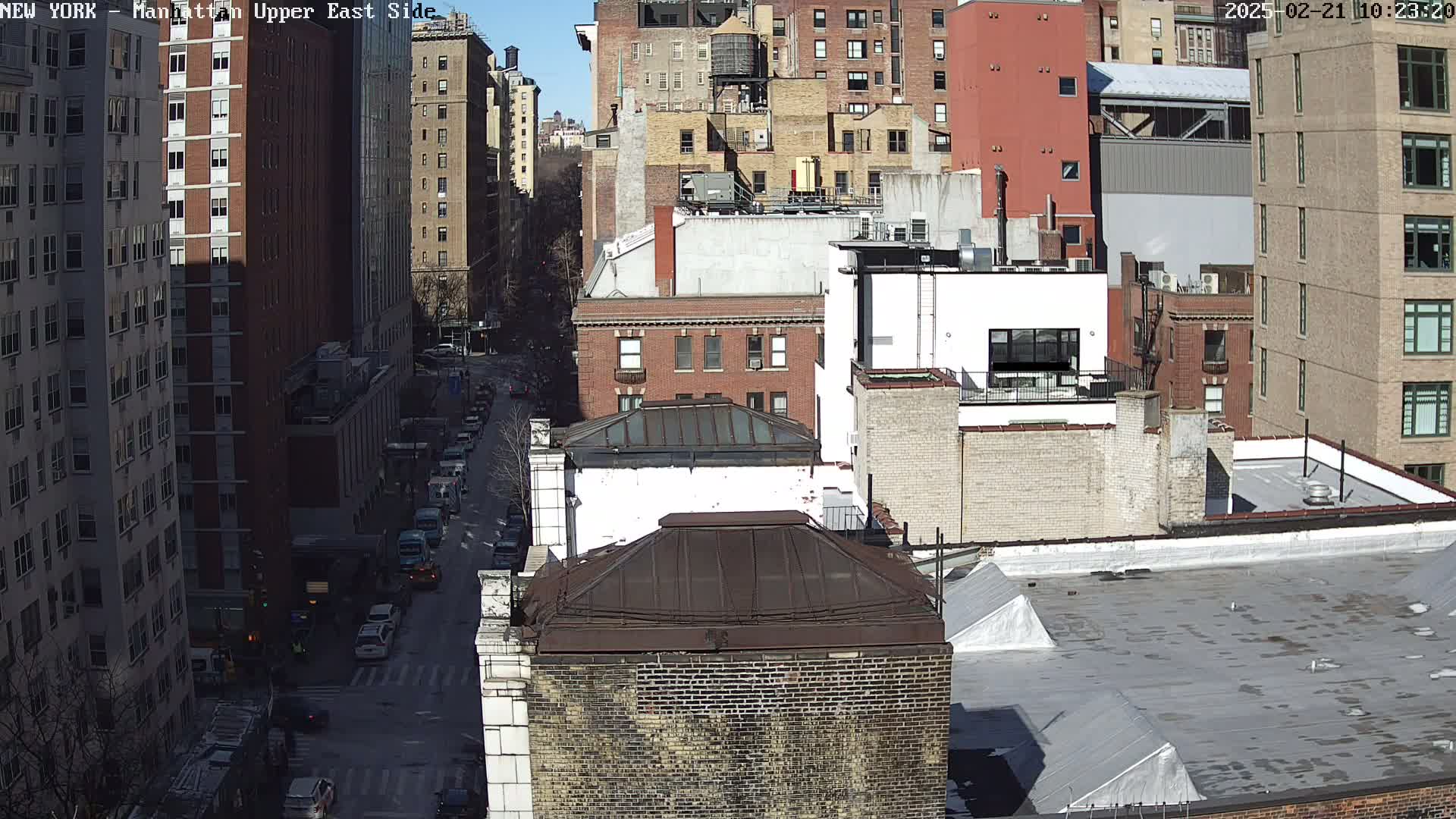 A sunny cityscape view of a Manhattan street, showing numerous brick buildings and a road with cars.