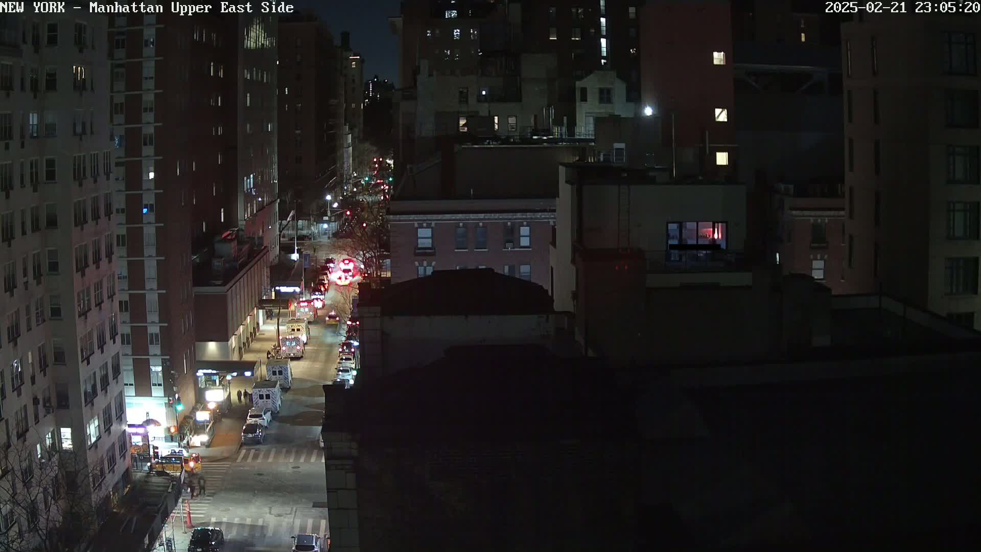 A nighttime aerial view shows city streets with multiple emergency vehicles and some traffic, under a dark sky.