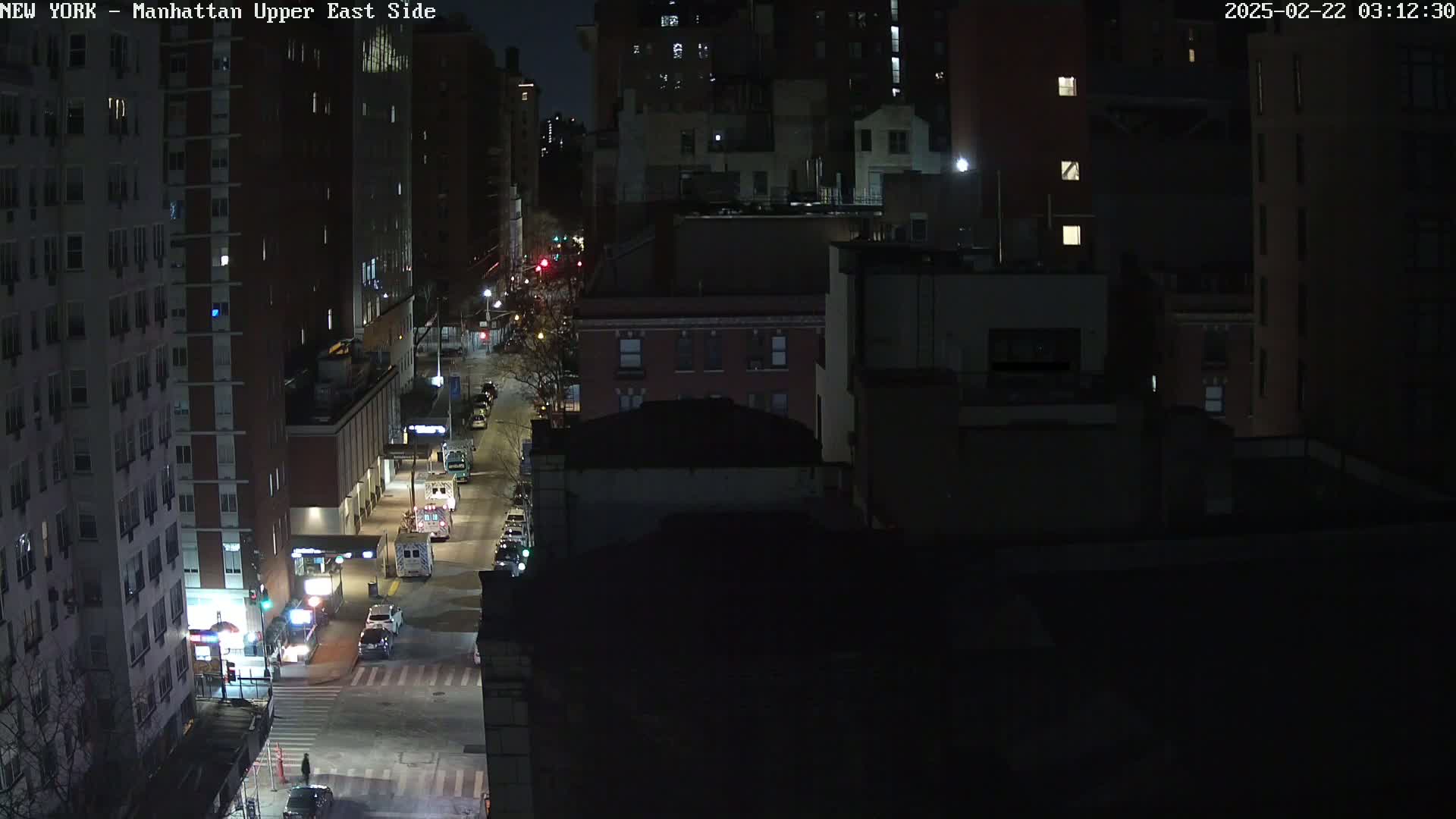 A nighttime, high-angle view of a city street with several vehicles, including what appears to be an ambulance, parked alongside buildings.