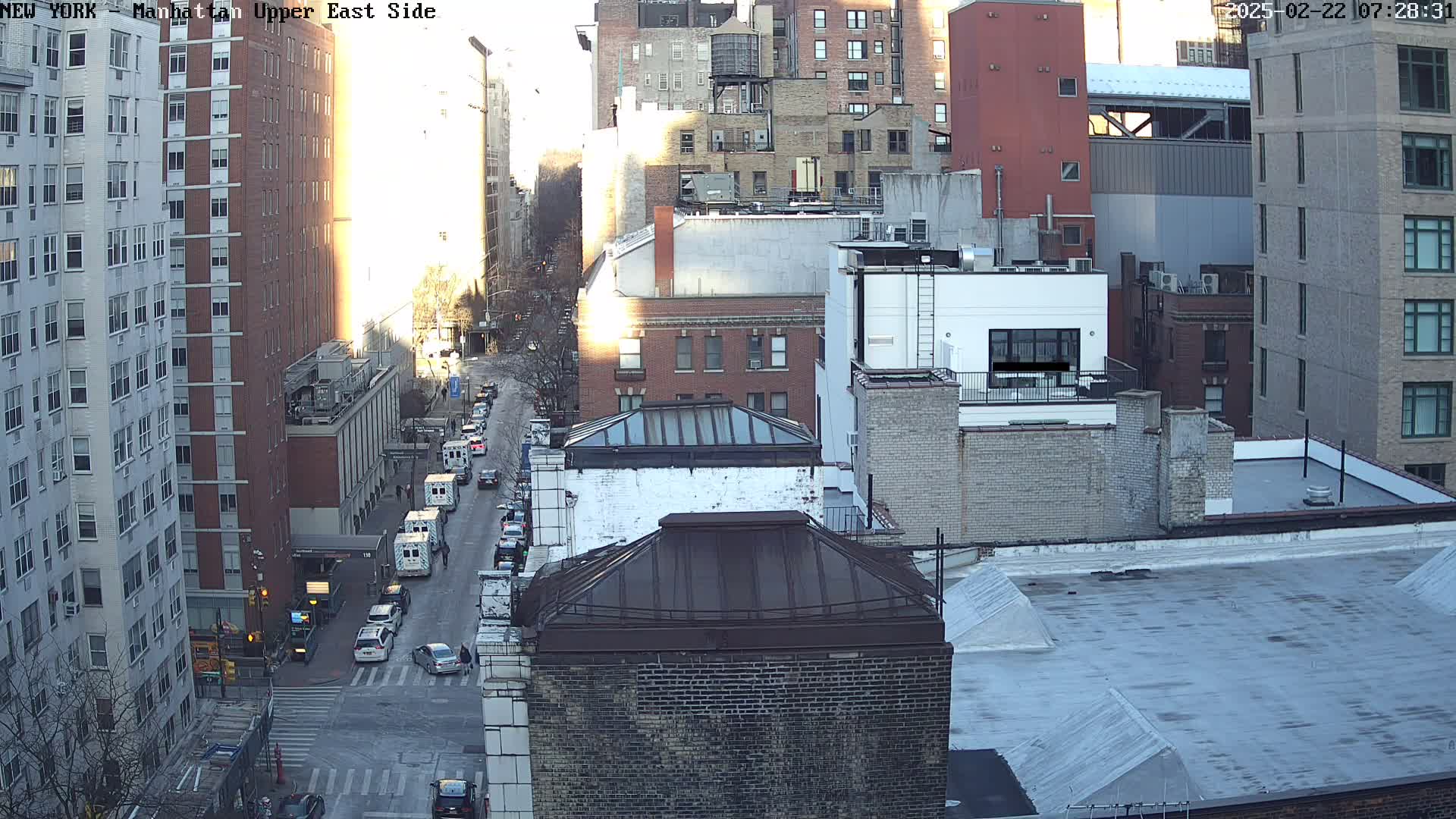 A sunny, city street scene viewed from above shows several multi-story buildings and a street with cars, including what appears to be an ambulance, at an intersection.