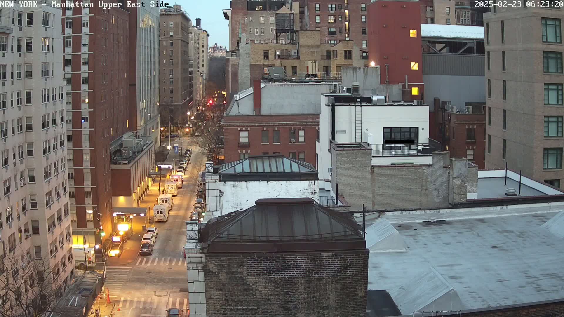 A twilight view of a city street shows several multi-story buildings, some vehicles parked and driving, and a few streetlights illuminating the scene.