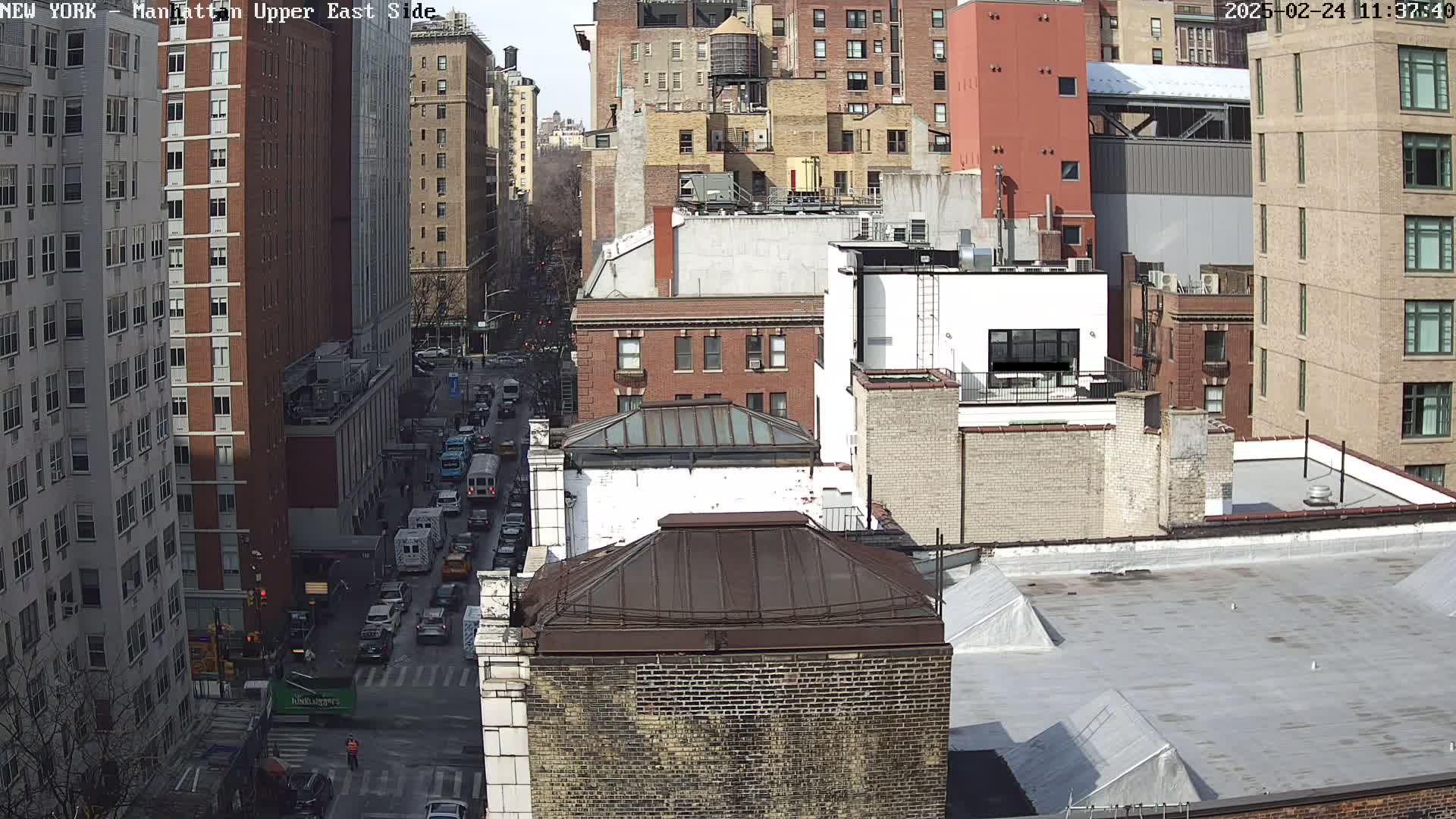 A sunny day reveals a high-angle view of a city street with various brick buildings, a few cars, and a pedestrian.