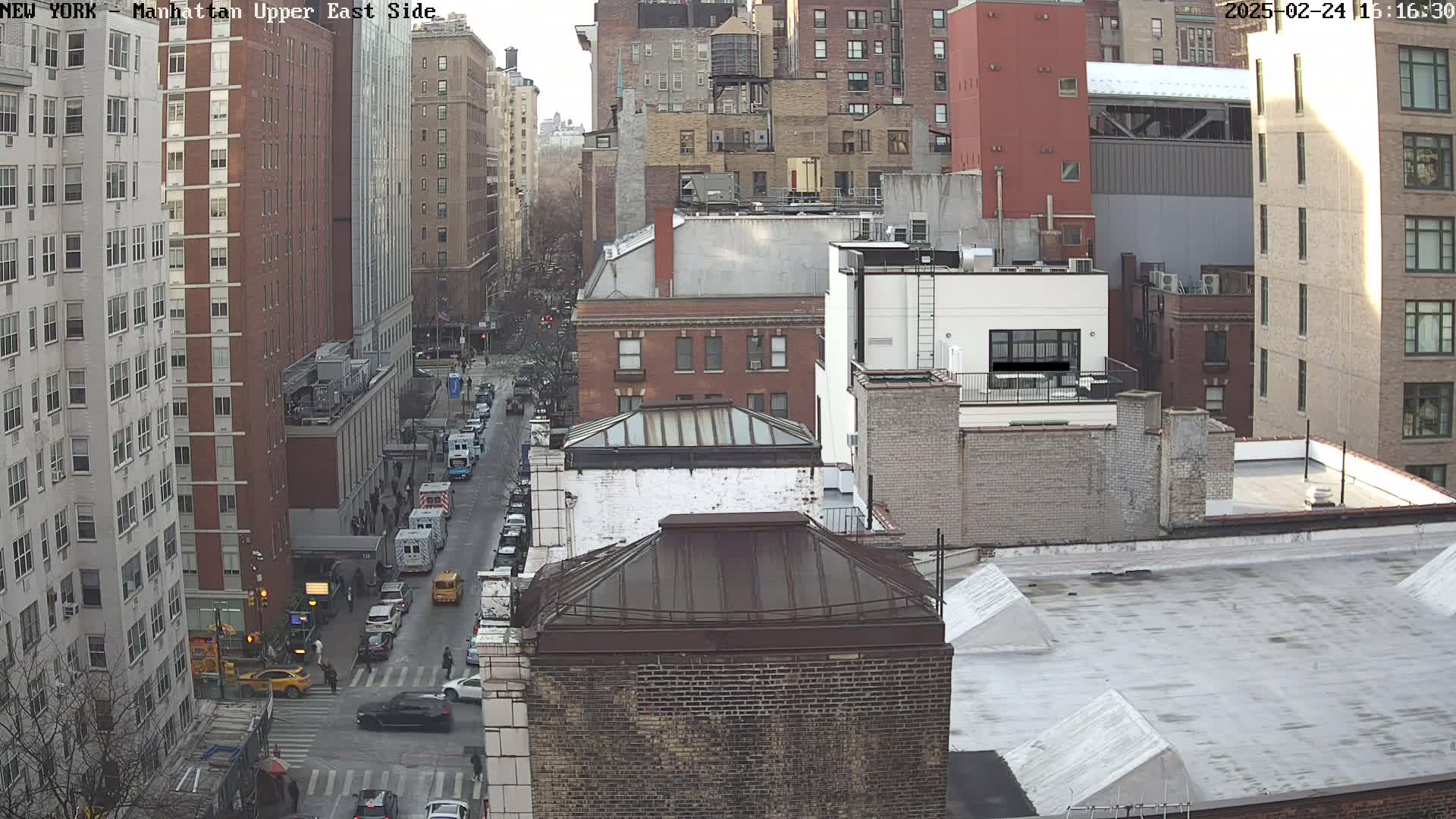 A high-angle view of a city street shows several multi-story buildings on a partly sunny day.