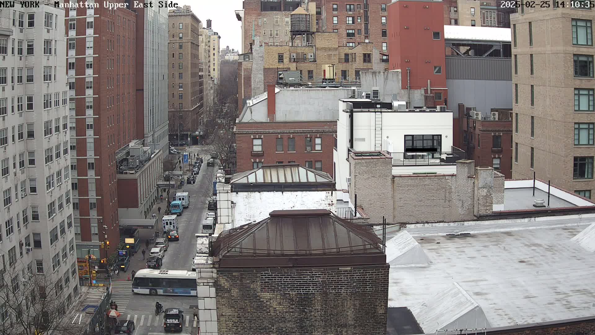 A high-angle view of a city street scene on an overcast day, showing multi-story brick buildings, vehicles on the road, and some bare trees.