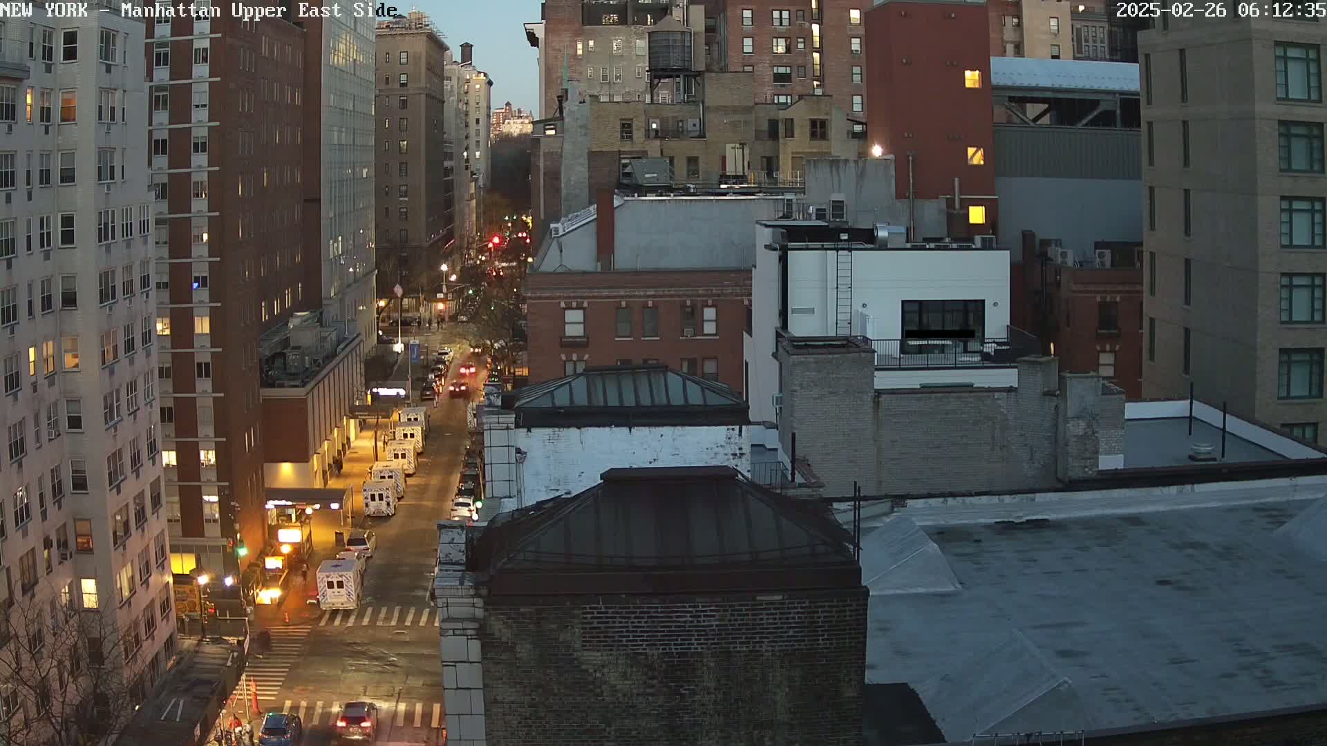 A twilight city street scene shows several tall buildings and vehicles, with a few emergency vehicles on the street.