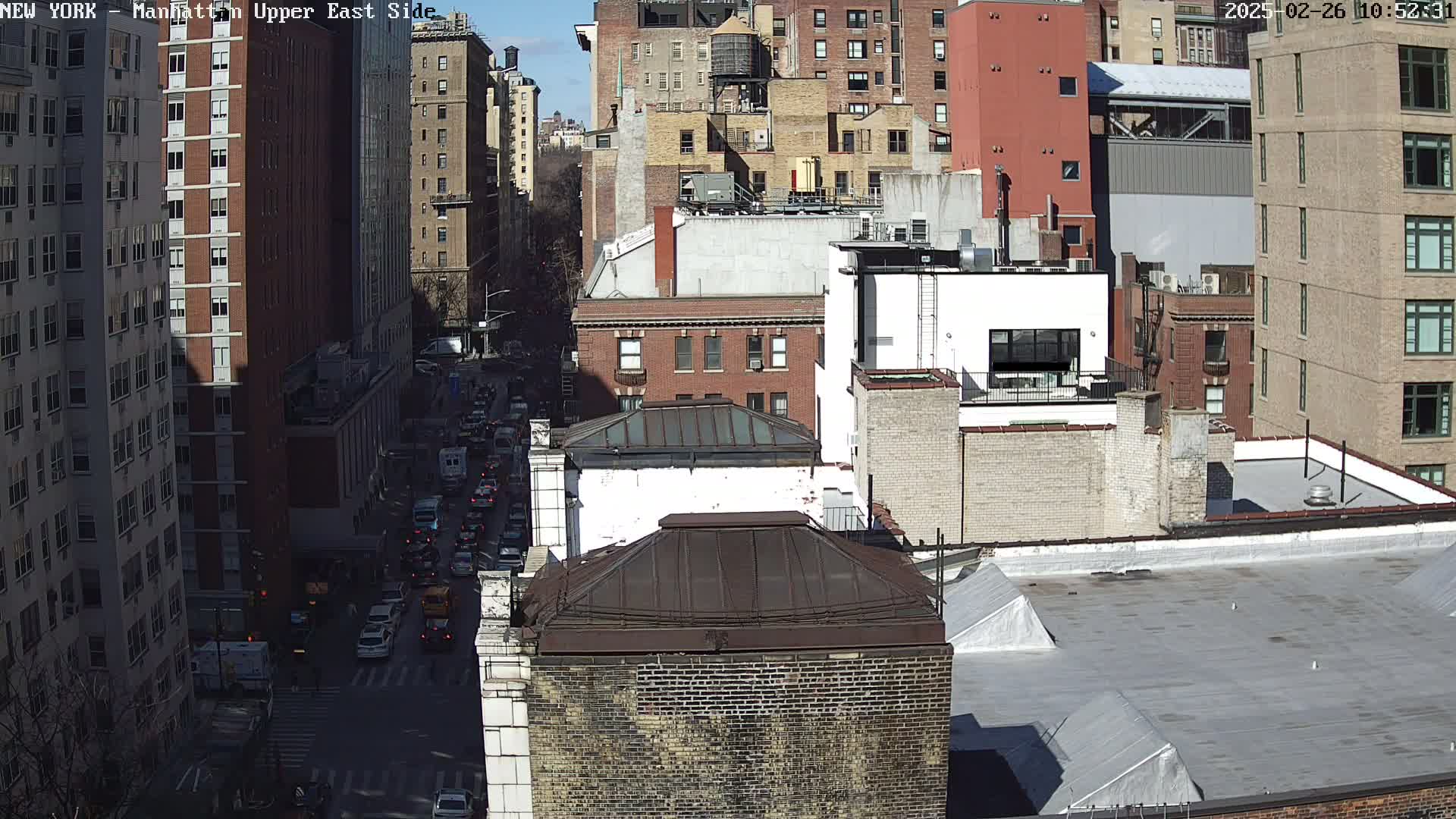 A sunny day view of a city street with many multi-story buildings and cars driving on a roadway below.