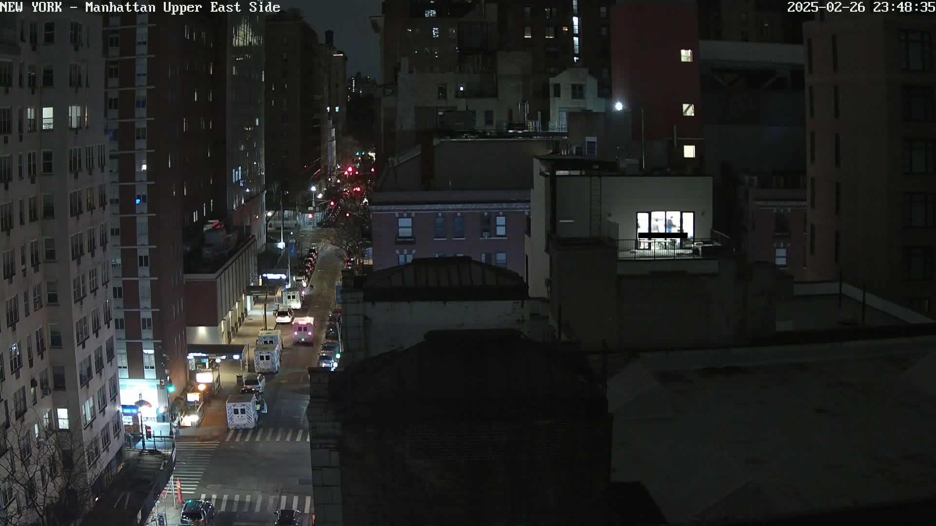 A nighttime aerial view of city streets and tall buildings, showing a few vehicles on a dimly lit road.