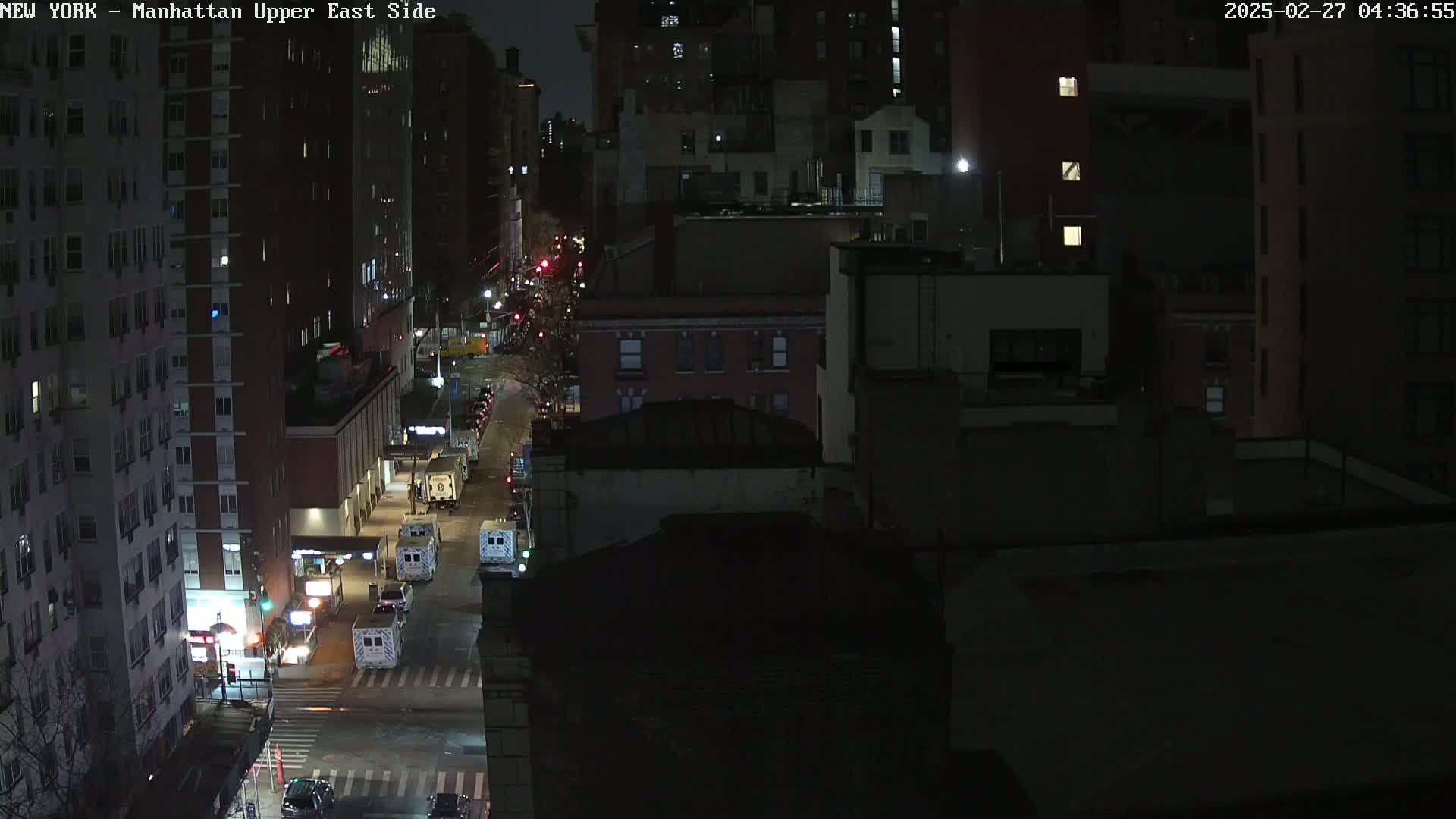 A nighttime aerial view shows a city street with several vehicles, including what appear to be emergency vehicles, parked along the side of the road in front of multi-story buildings.