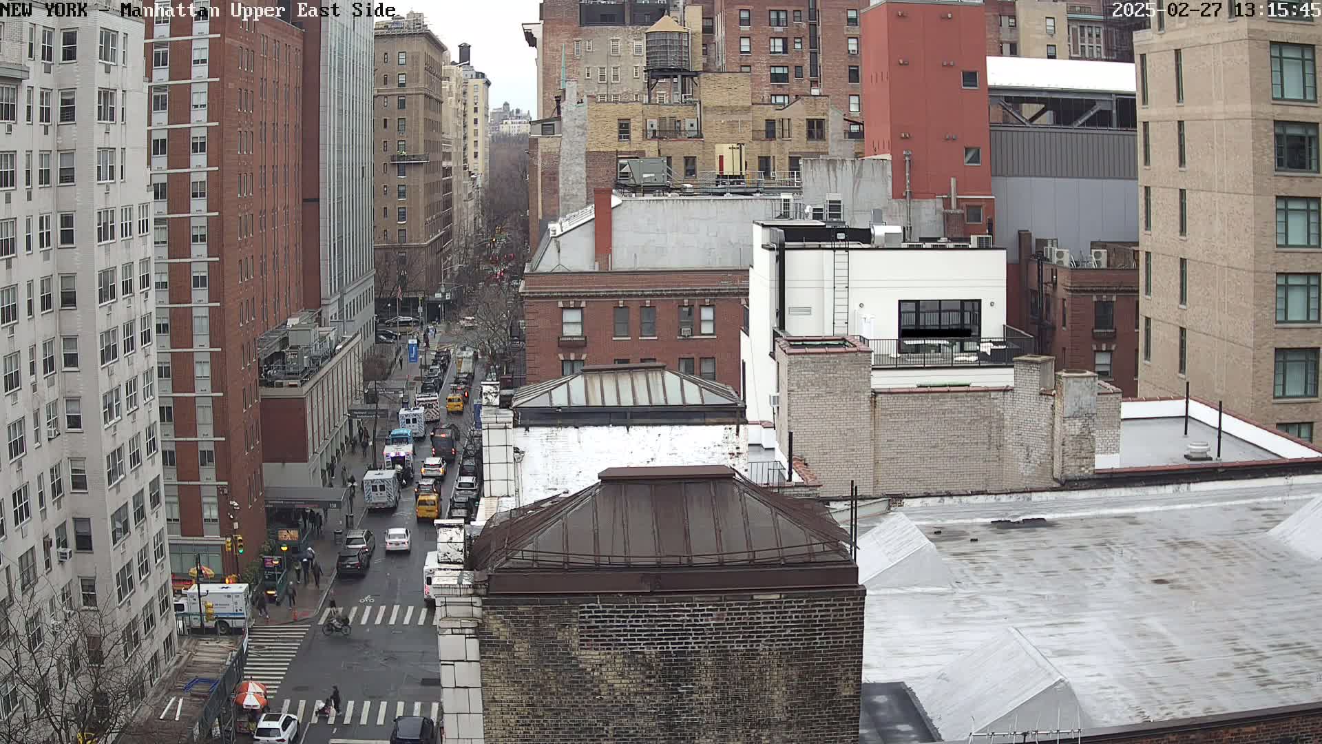 A high-angle view of a city street with traffic on a cloudy day, showing multiple brick buildings and a water tower.