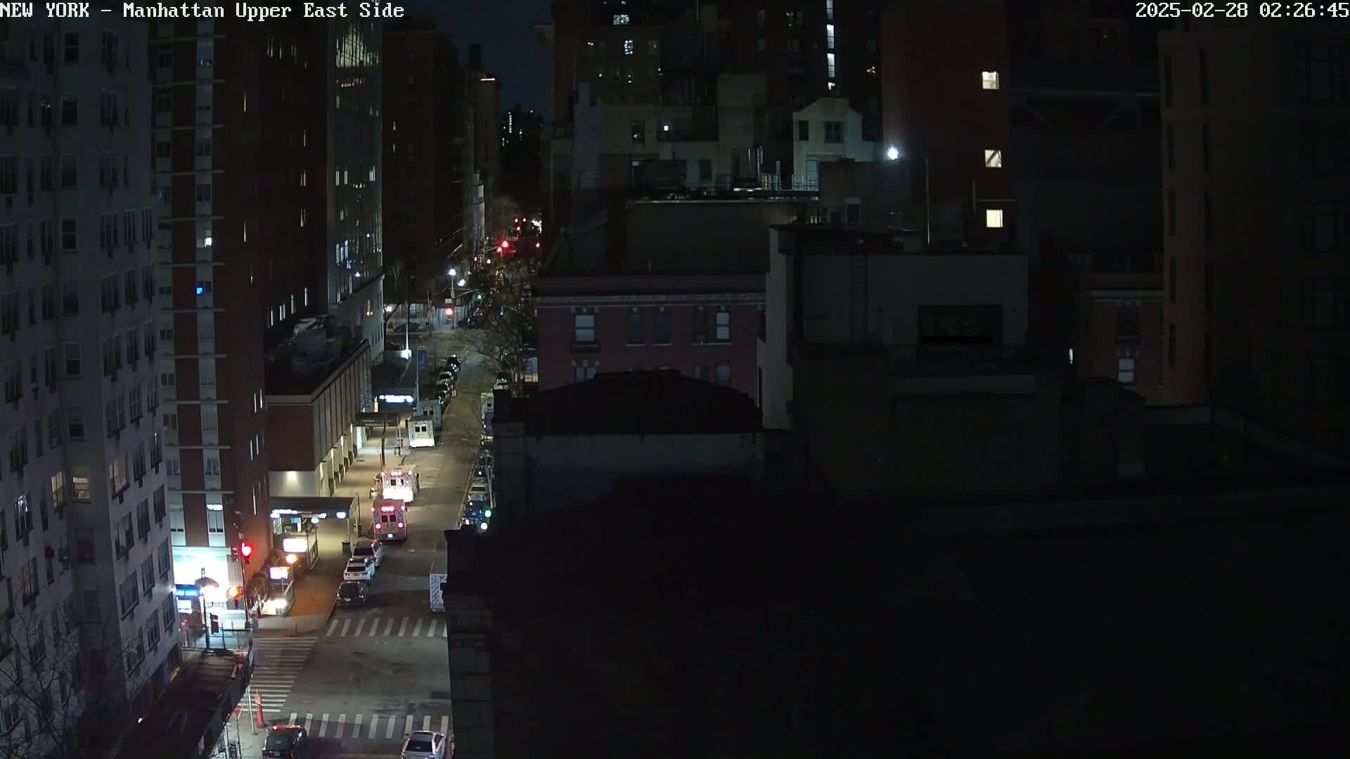 A nighttime, high-angle view of a city street with several vehicles, including an emergency vehicle, parked along the side of a multi-lane road, surrounded by dark buildings.