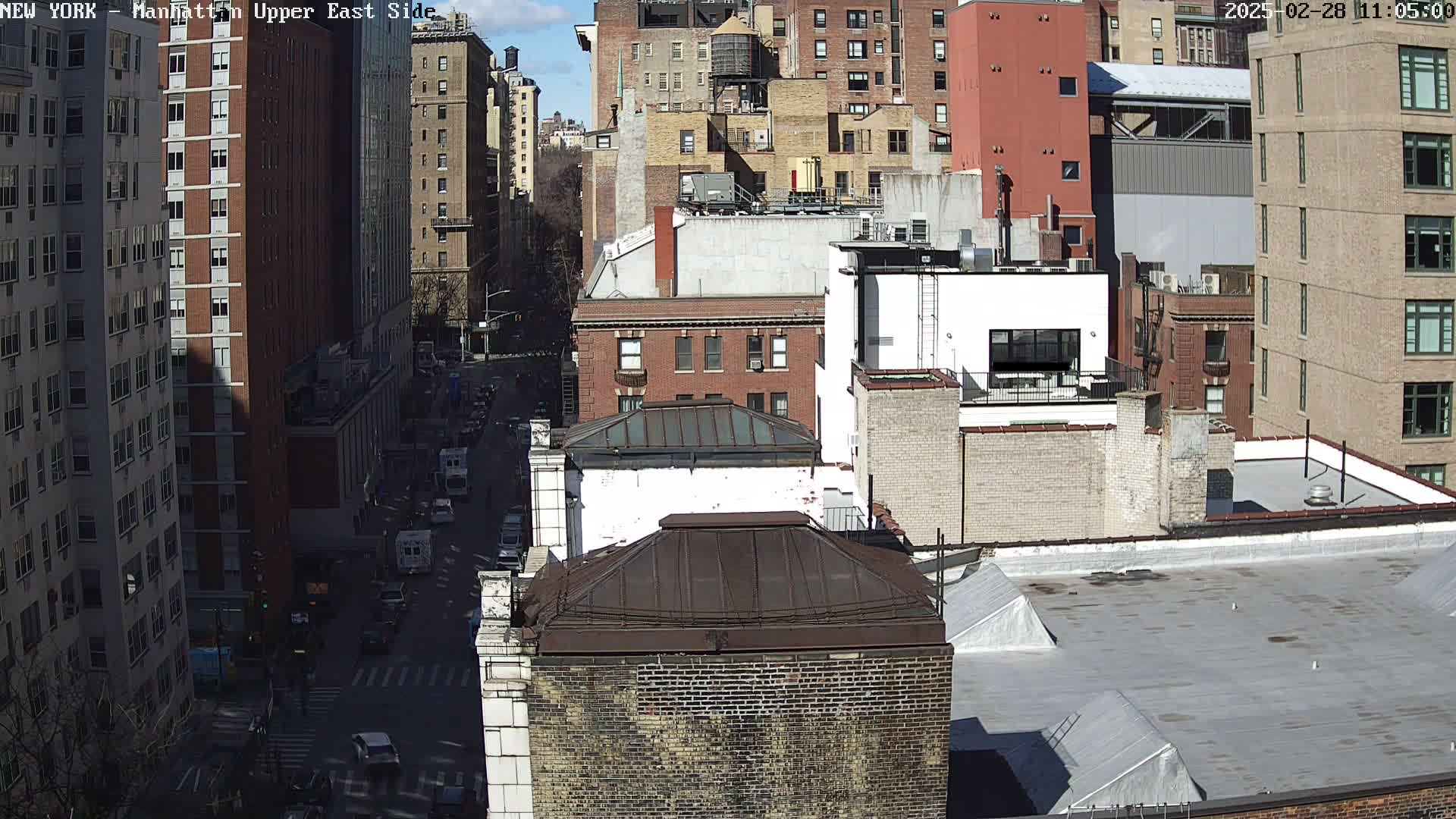 A sunny day view of a densely packed city with brick buildings, rooftops, and a street with cars below.