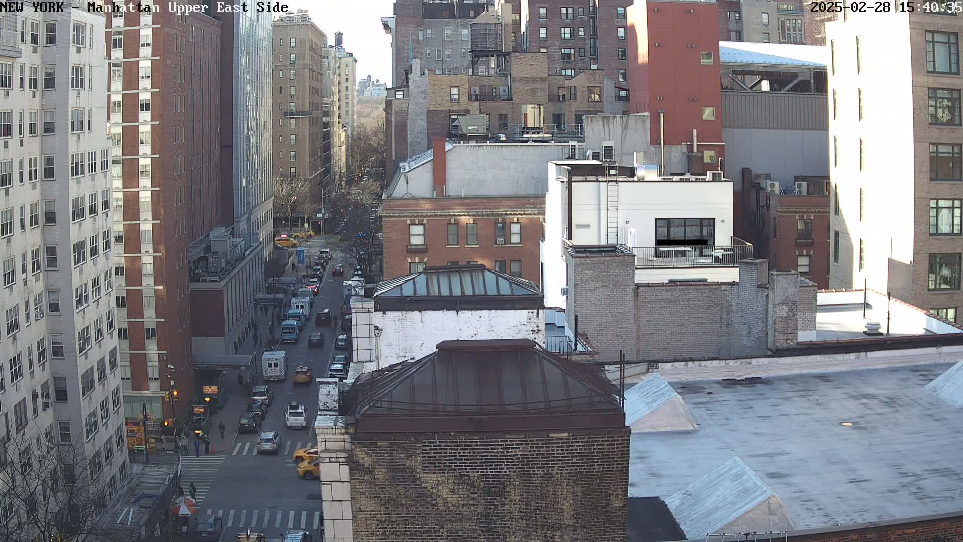 A sunny day view of a city street with traffic and numerous brick buildings, showing rooftops and street-level activity.