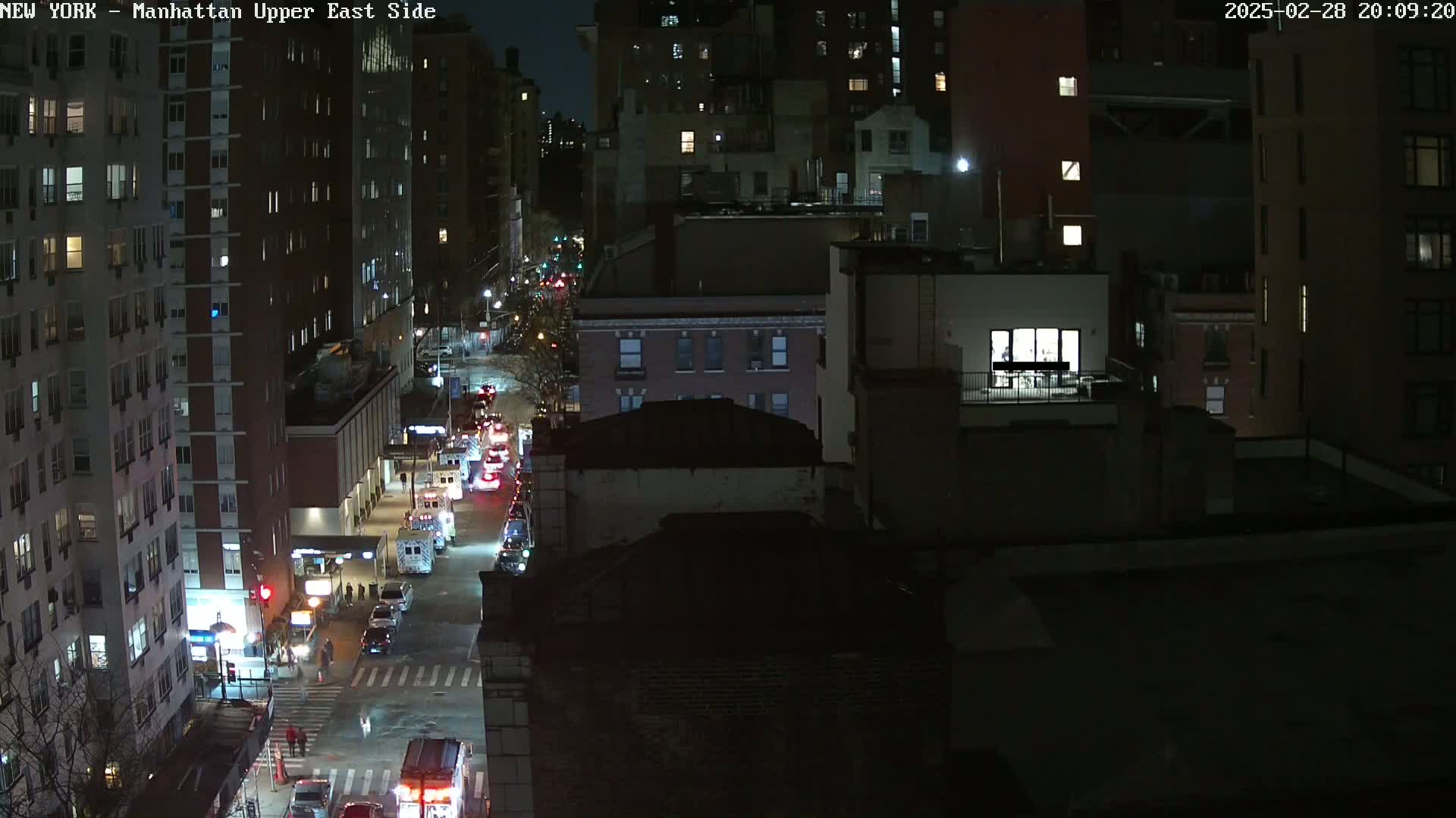 A nighttime aerial view of a city street with several buildings, some vehicles, and pedestrians.