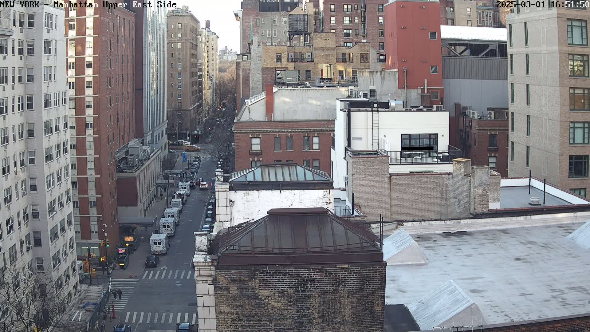 A high-angle, long shot of a city street, with several multi-story brick buildings and a few parked vehicles under a mostly cloudy sky.
