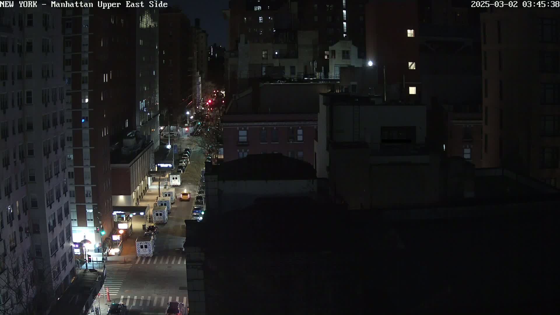 A nighttime aerial view shows city streets with several vehicles, including what appear to be emergency vehicles, parked along the sides of dark city buildings.