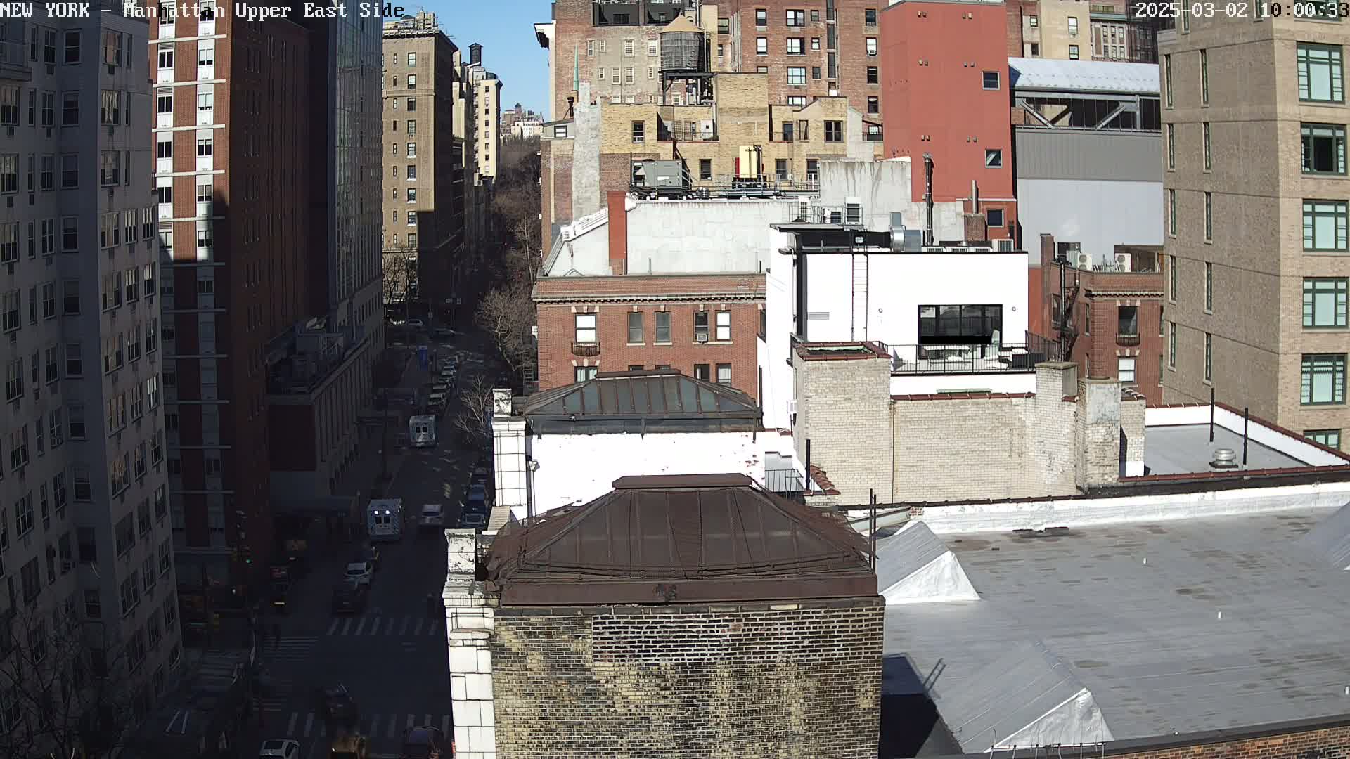 A sunny cityscape view of numerous brick buildings, some with rooftop structures, and a street with sparse traffic below.